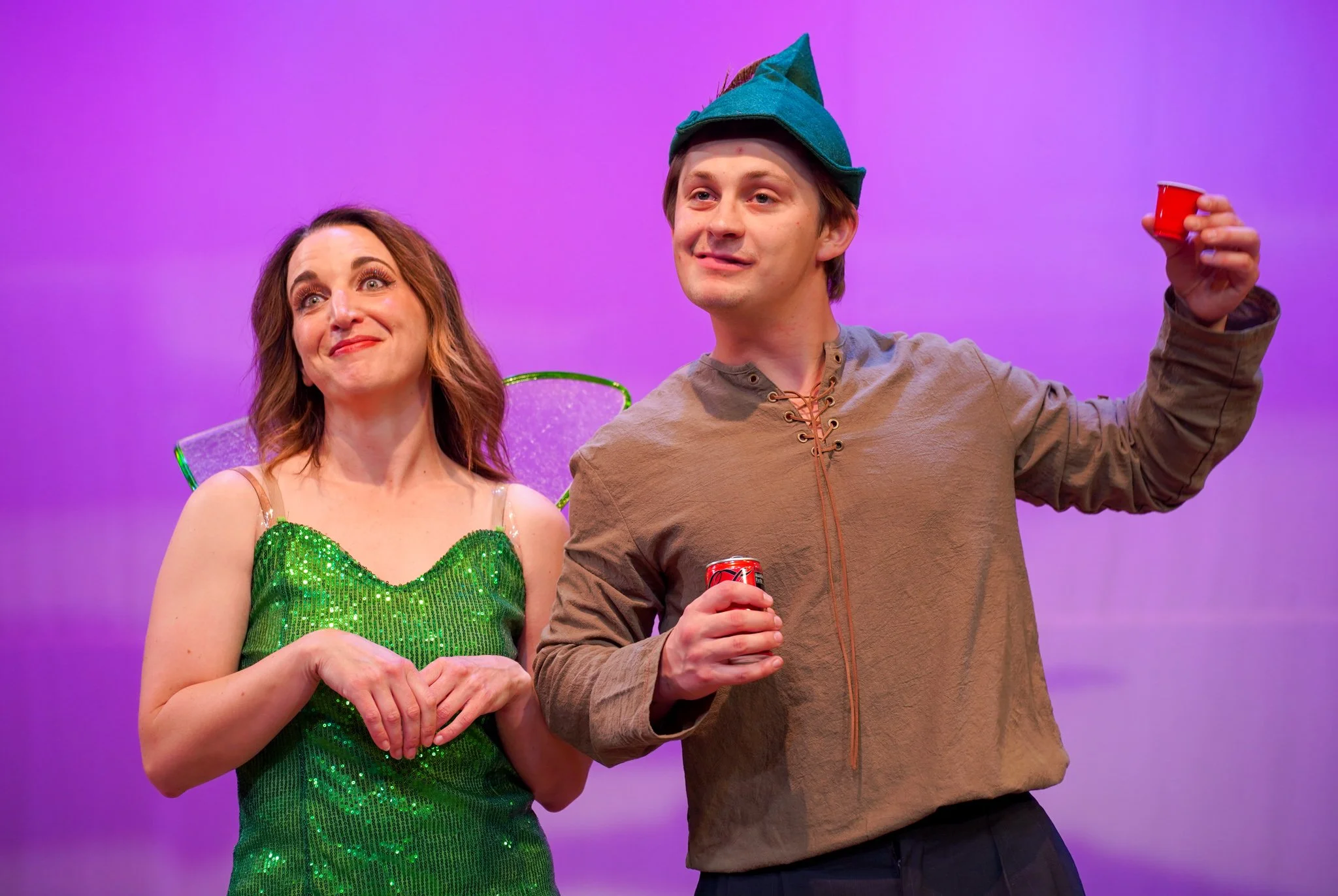 Woman in a glittery green dress and a man in a tan shirt with a green hat, both holding small red cups, standing on a purple background.