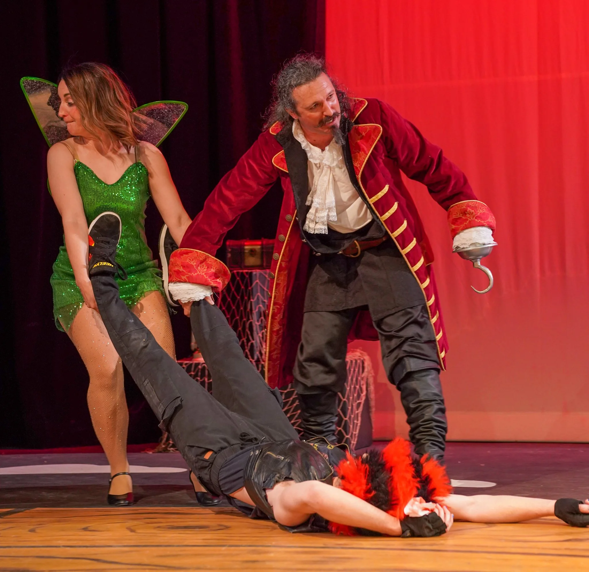 Two performers on stage, one lying on the floor and the other kneeling beside them, dressed in elaborate costumes, with the background featuring a red curtain and stage props.