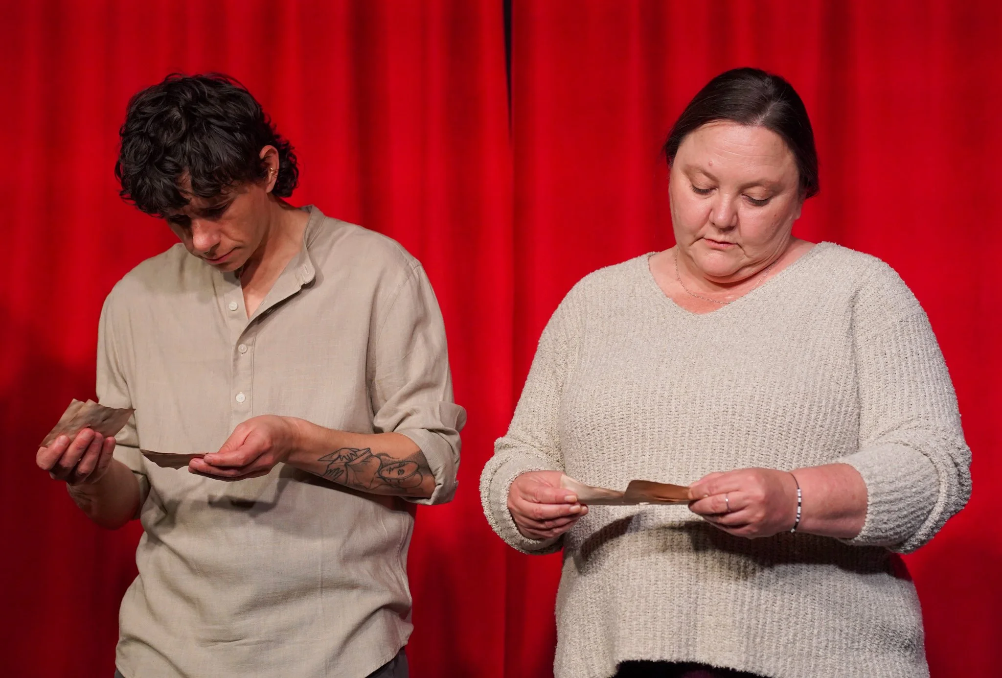 Two women standing on stage in front of red curtains, looking down at papers in their hands.