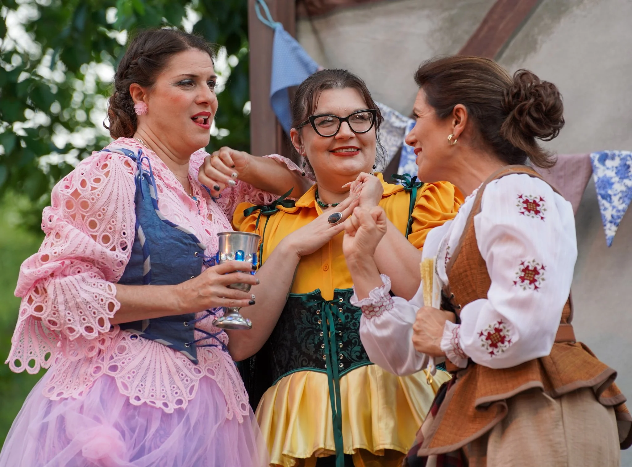 Three women dressed in traditional clothing sharing a moment at an outdoor event with decorations behind them.