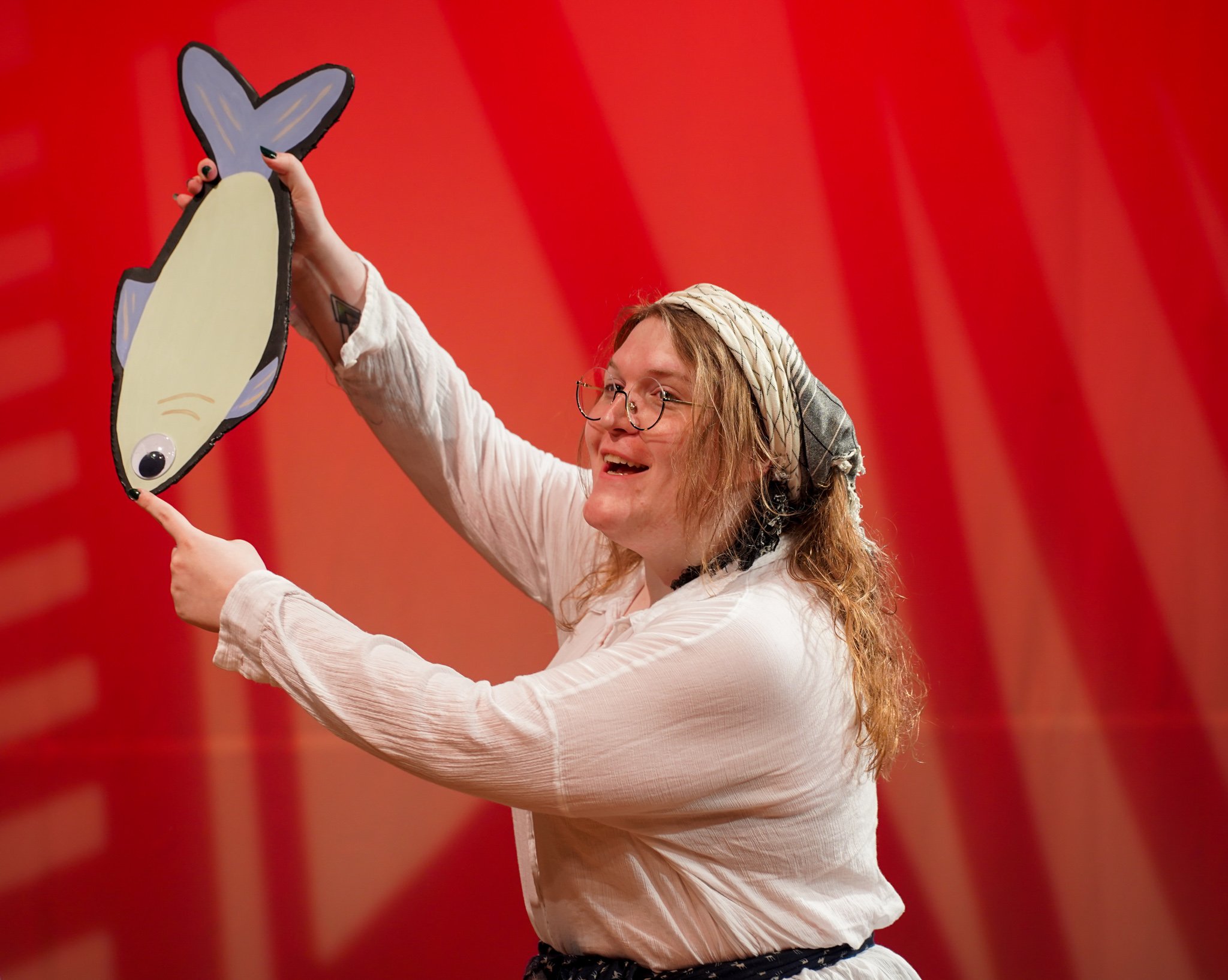 A woman with long red hair, glasses, and a headscarf holding a large, cartoon-style fish puppet with a red background.