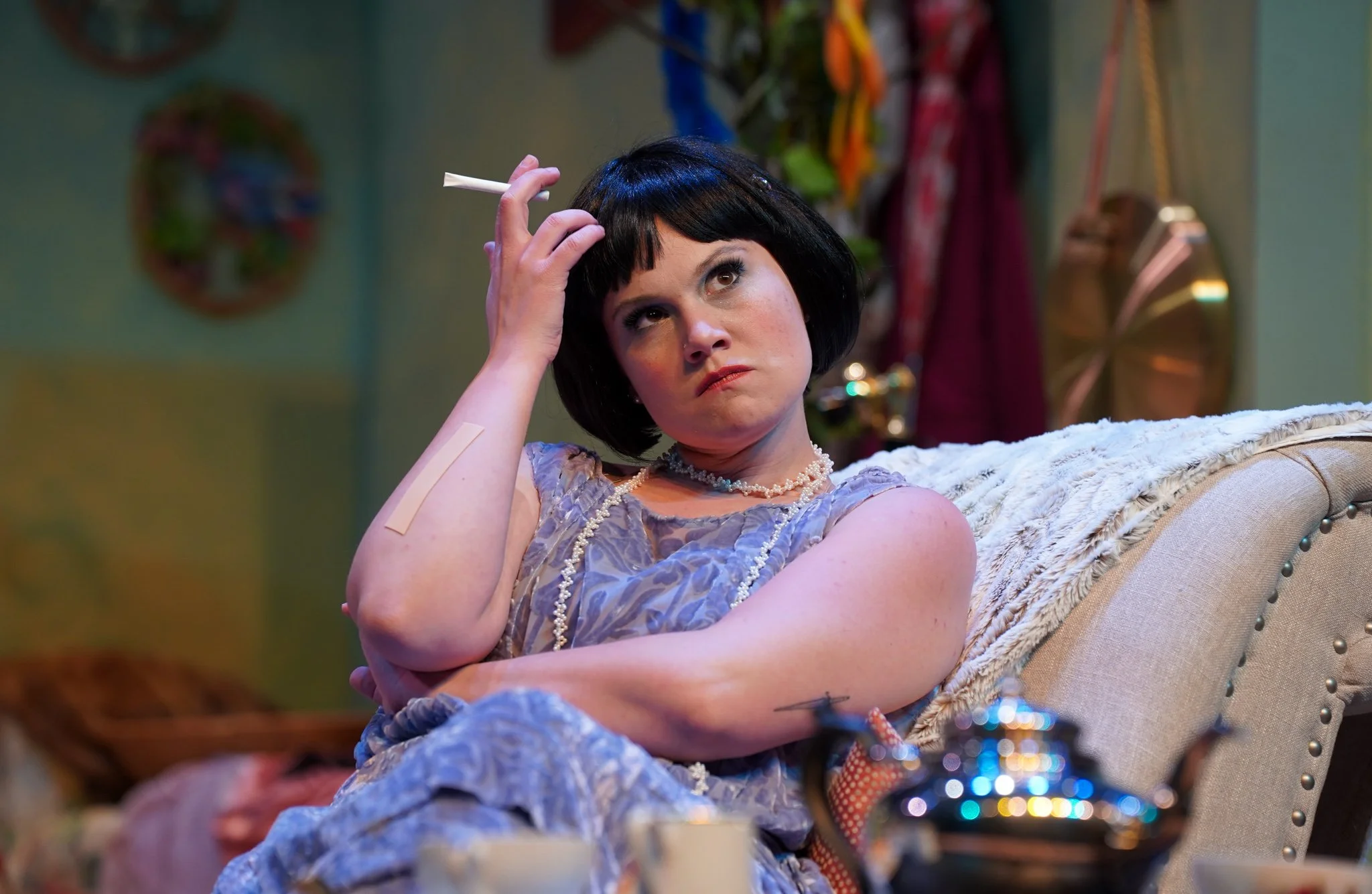 A woman with short black hair and a pearl necklace sitting on a vintage cream-colored sofa, holding a cigarette in her right hand, looking up with a pensive expression, in a room with colorful wall art and hanging decorations.