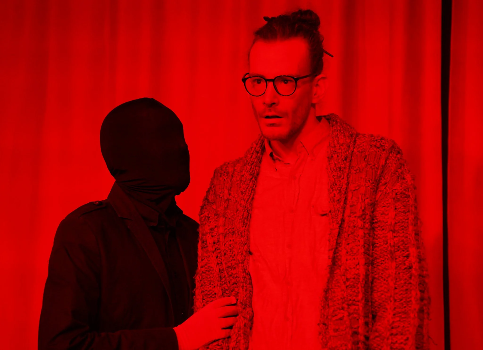 A man with glasses and a woman with a head covering standing and talking in front of a red curtain.