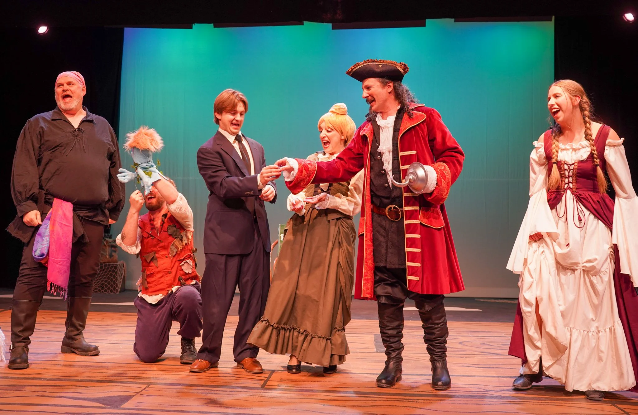 Actors on stage performing in a theatrical production, dressed in costumes that resemble pirates or historical characters, with some pointing and laughing, set with a plain colored backdrop.