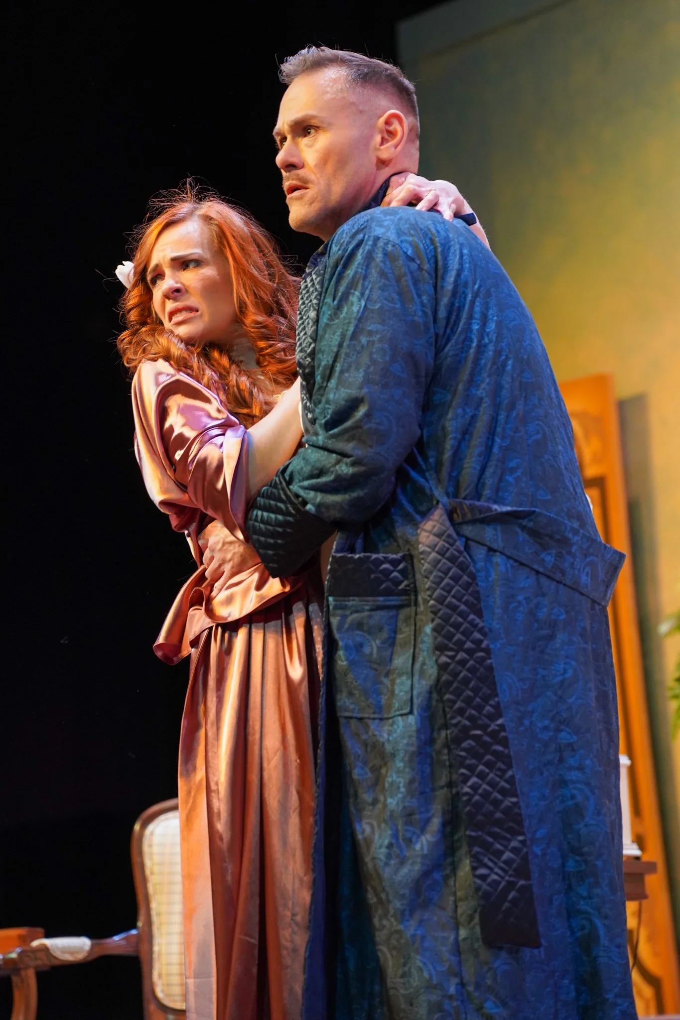 A man and a woman are on stage in a theatrical performance, showing intense emotional expressions. The woman has red hair, wears a peach-colored dress, and appears distressed. The man has short hair, is dressed in a dark blue patterned robe, and look