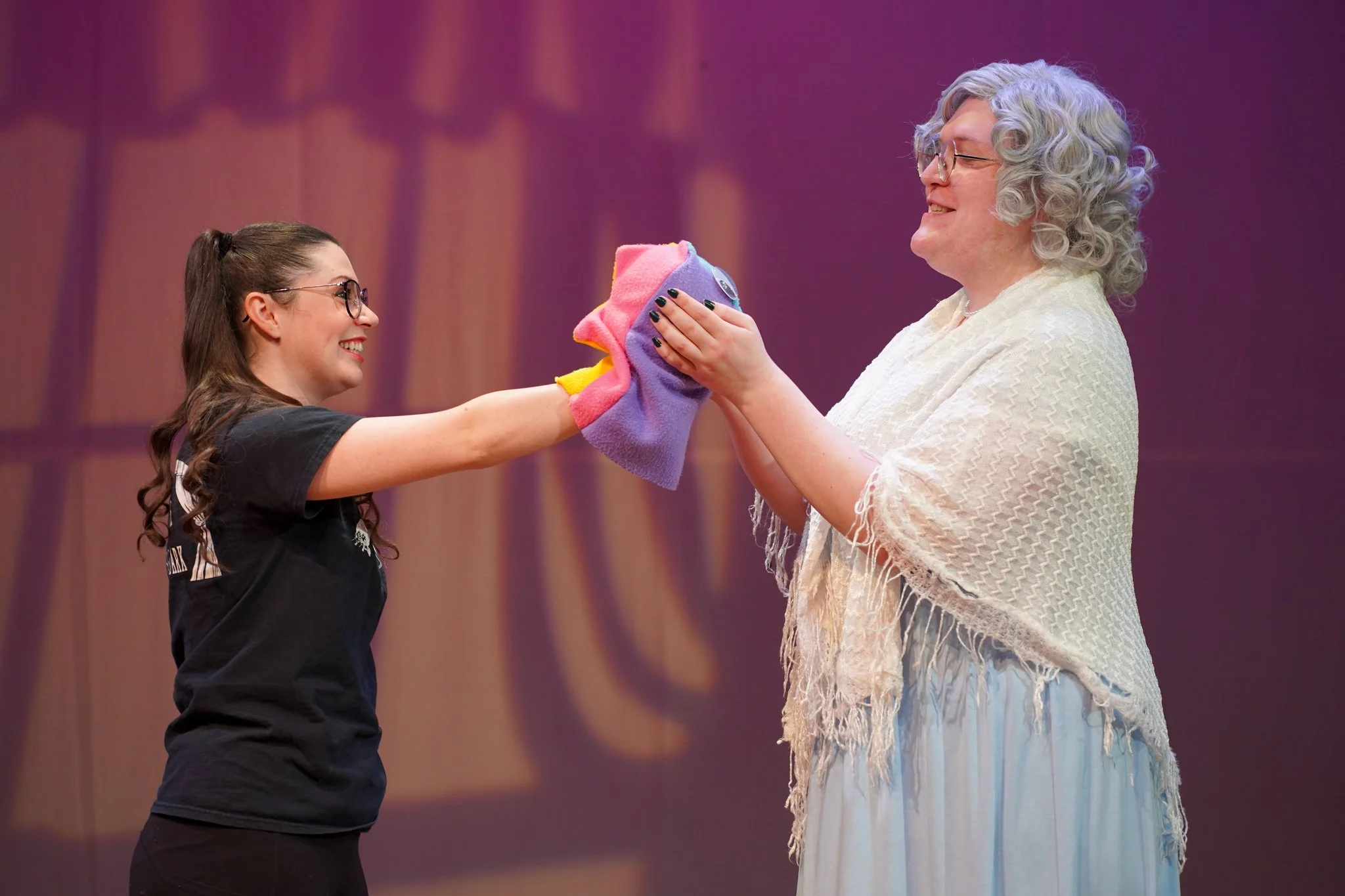 A young woman with glasses and dark hair tied in a ponytail is smiling and holding a colorful cloth, offering it to an older woman with curly gray hair who is also smiling. The older woman is wearing a white, fringed shawl. They are on a stage with a