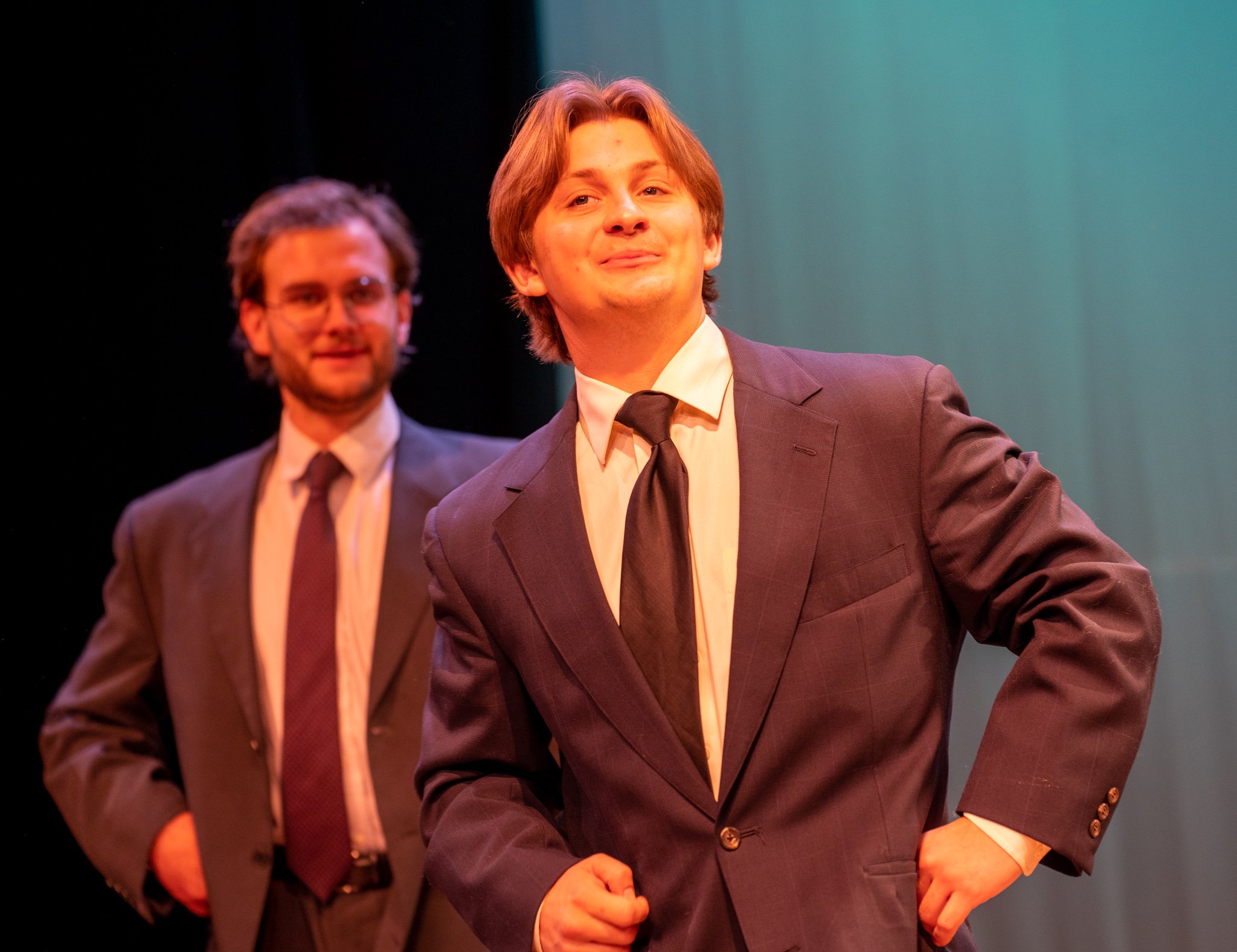 Two men are dressed in suits and ties, standing on a stage with a dark background and a gradient green curtain. The man in front is smiling with a confident expression, and the man in the background has glasses and is slightly out of focus.