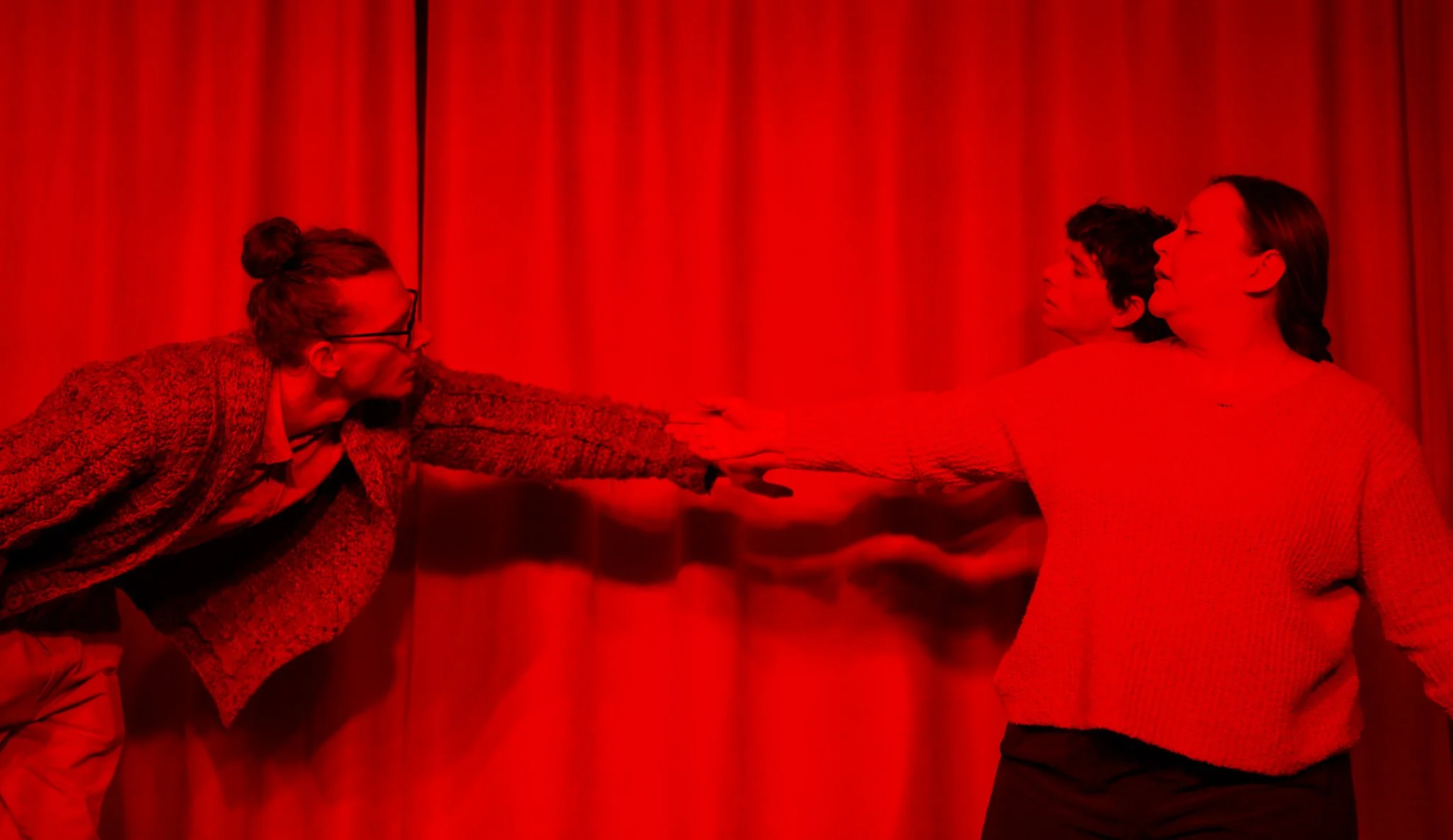 Three women, all with dark hair, are standing in front of red curtains. Two women are facing each other, with one woman holding the other's hand, creating a visual of a handshake or connection. The women are dressed casually.