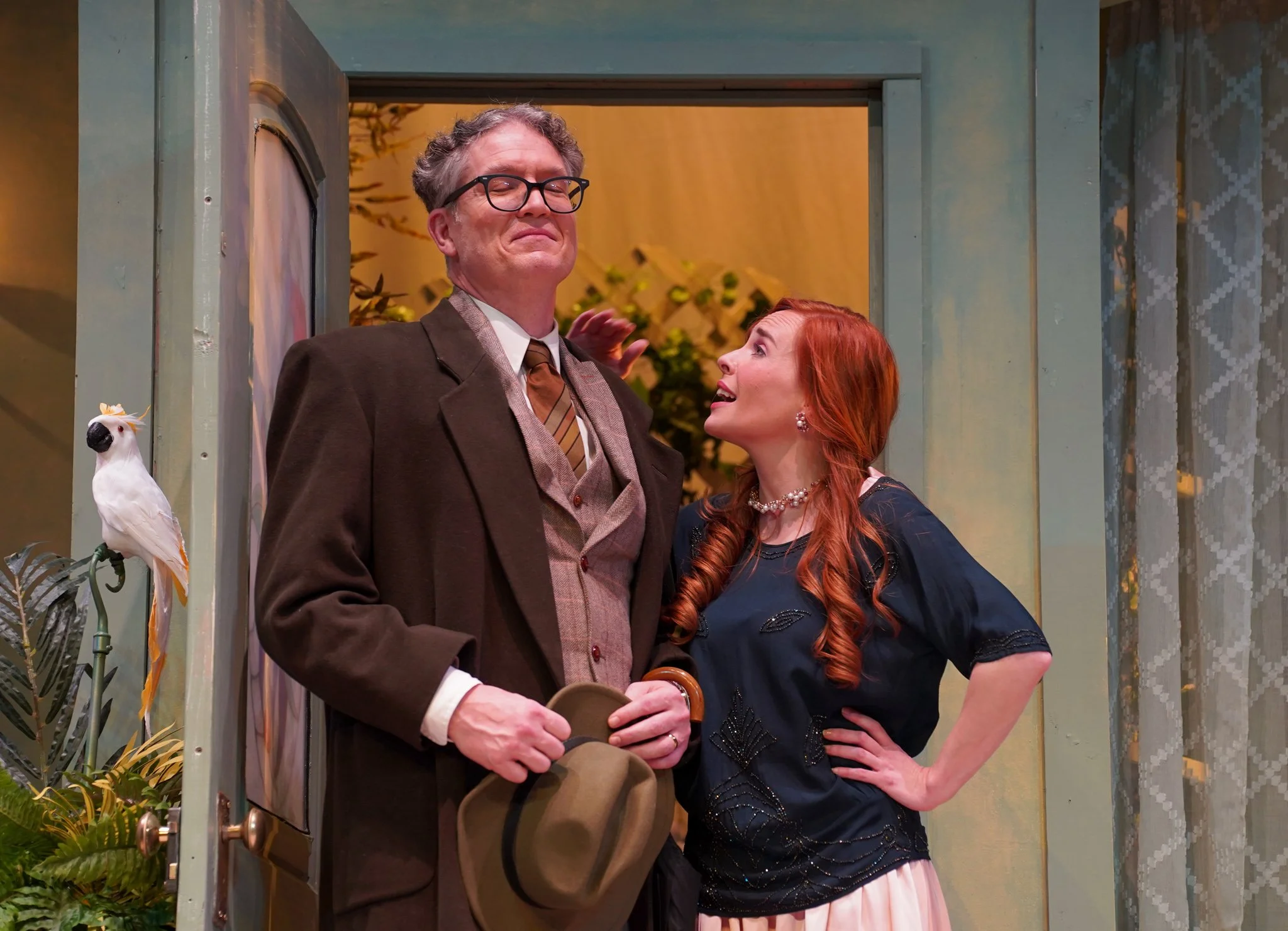 A man with glasses and a woman with red hair and pearl jewelry stand close together at an event, smiling. The man holds a fedora hat and a cane, and appears to be emotional. The woman is gesturing with her right hand and gazing at the man. They are i