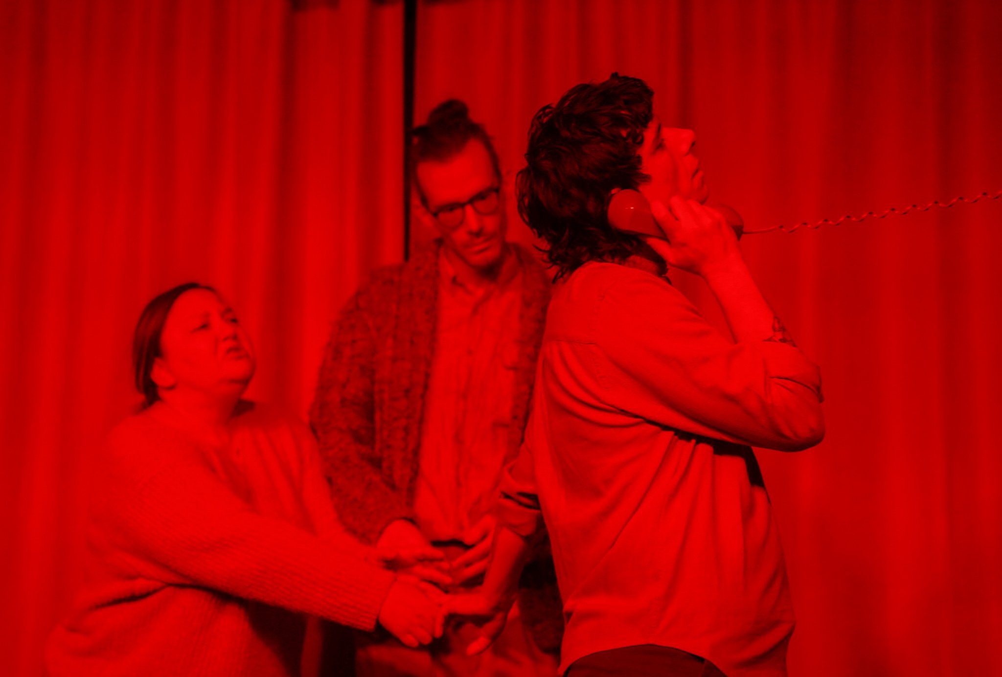 People standing in front of red curtains, with a woman on the right talking on a telephone and others looking ahead.