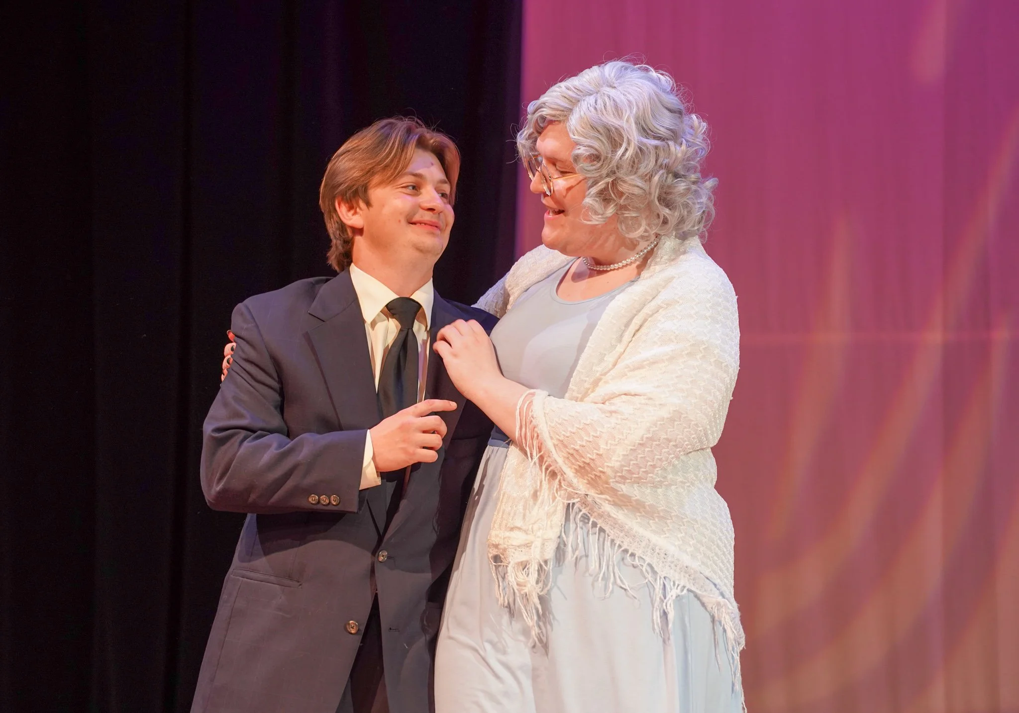 A young man in a suit and an older woman with curly gray hair and glasses are smiling and looking at each other in a warmly lit setting.