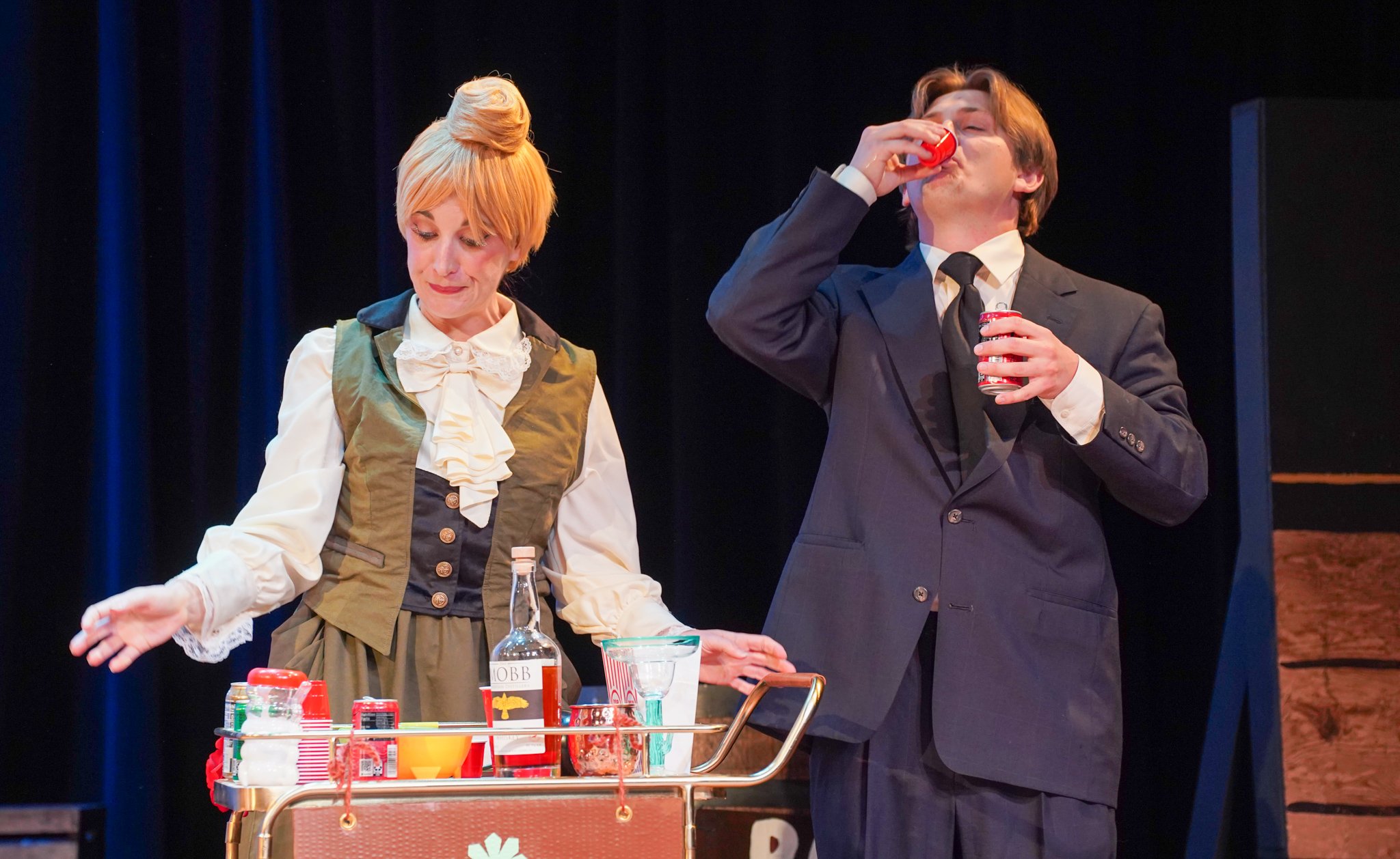 A scene from a stage play with two actors in vintage clothing. The woman with short blonde hair is standing behind a table with various condiments and a bottle of JOBB syrup, looking down with a slight smile. The man, in a dark suit, is drinking from
