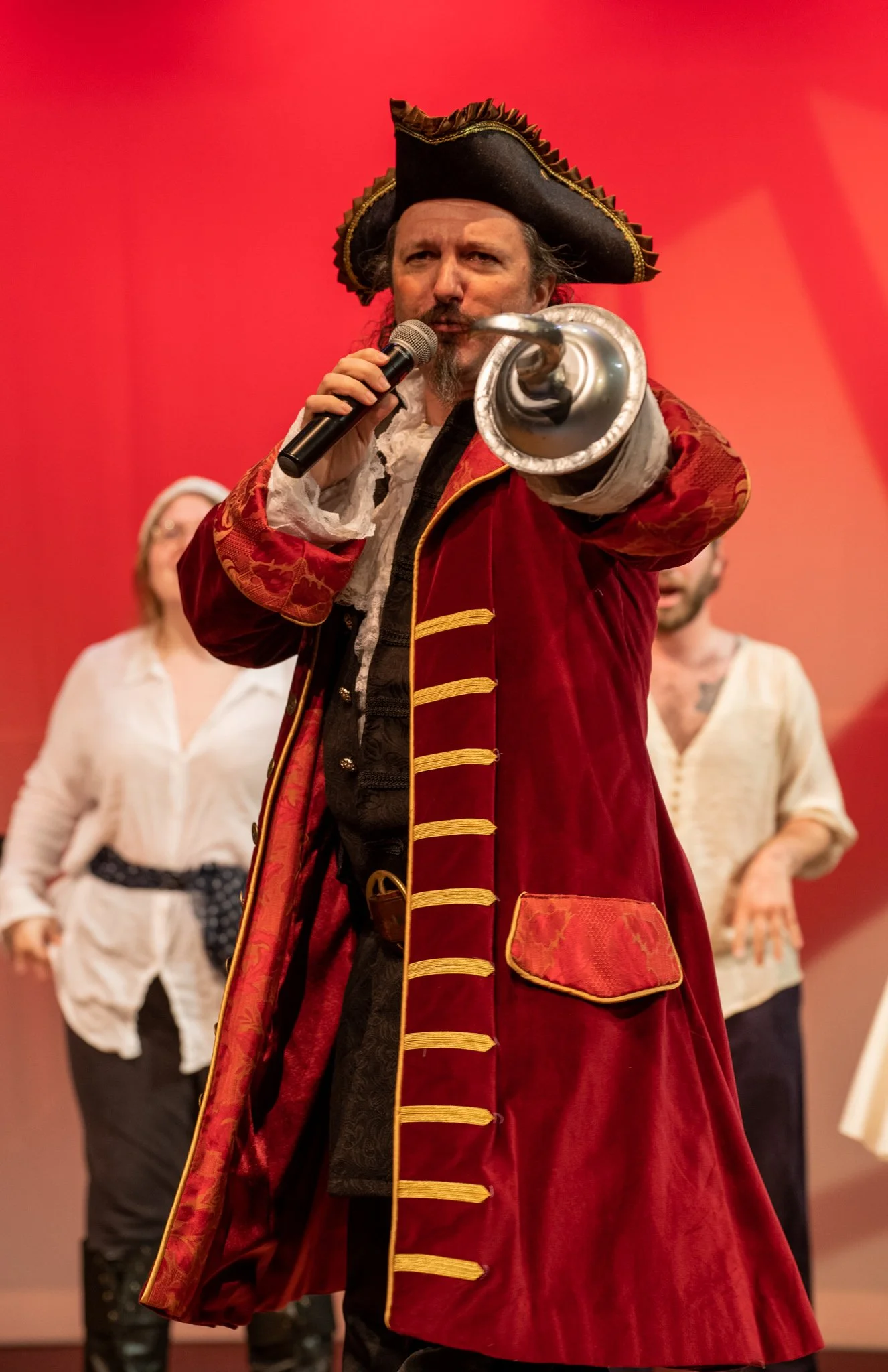 A man dressed as a pirate is pointing towards the camera, holding a small horn, with a red background. There are two women in the background, one in a white blouse and the other in a white shirt, blurred.