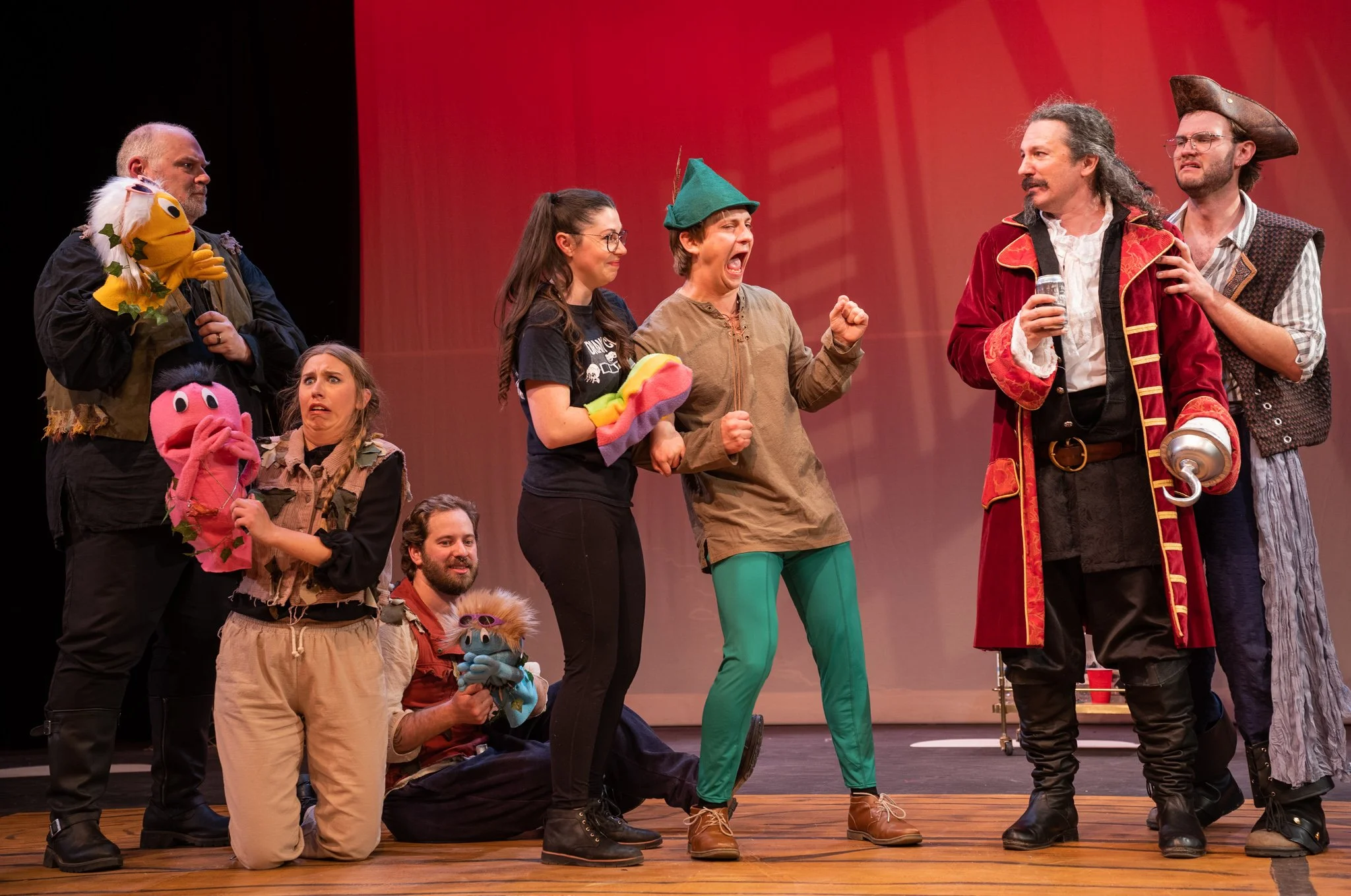 A group of actors dressed in costumes from 'Peter Pan' on stage, some holding puppet characters, standing on a wooden stage with a red backdrop.