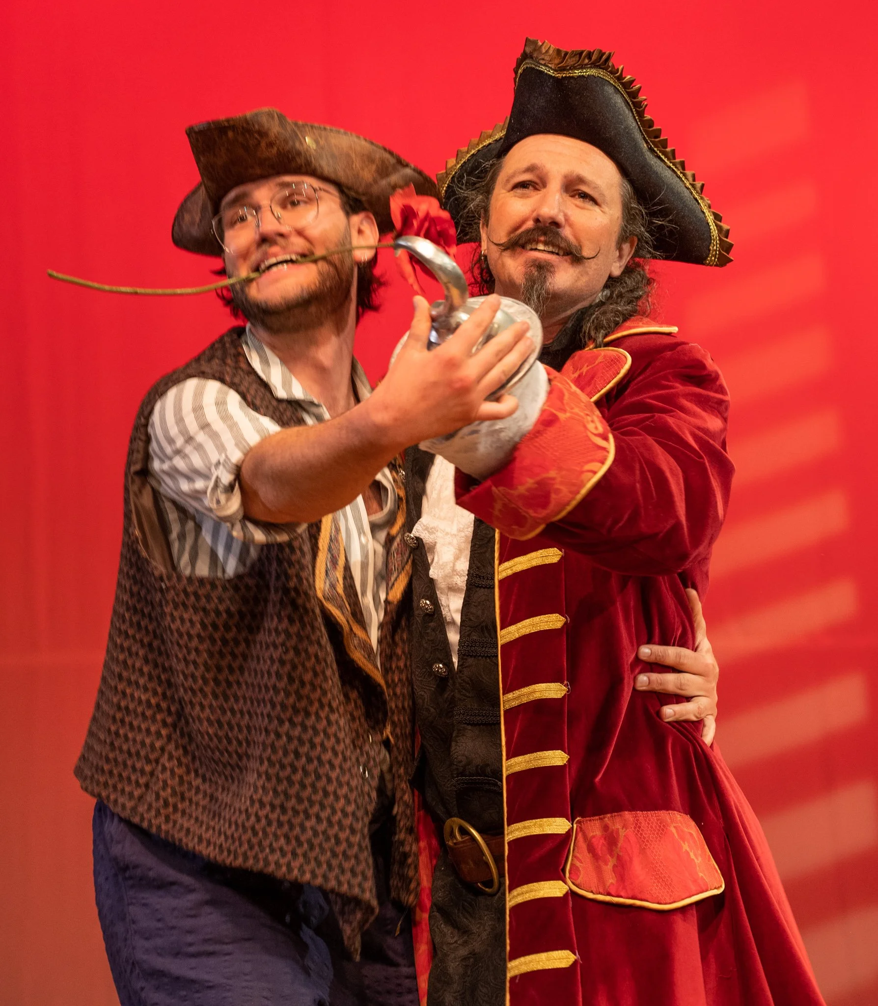 Two men dressed as pirates, with one holding a sword and a rose, standing close together against a red background.