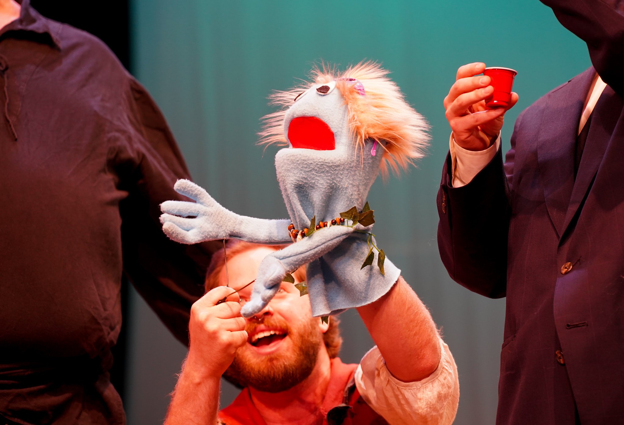 A puppet with a white face, orange hair, and a red tongue, being manipulated by a person with a beard wearing a beige shirt, while two other people, one in a dark suit and the other in a purple jacket, observe or participate in the puppet show.