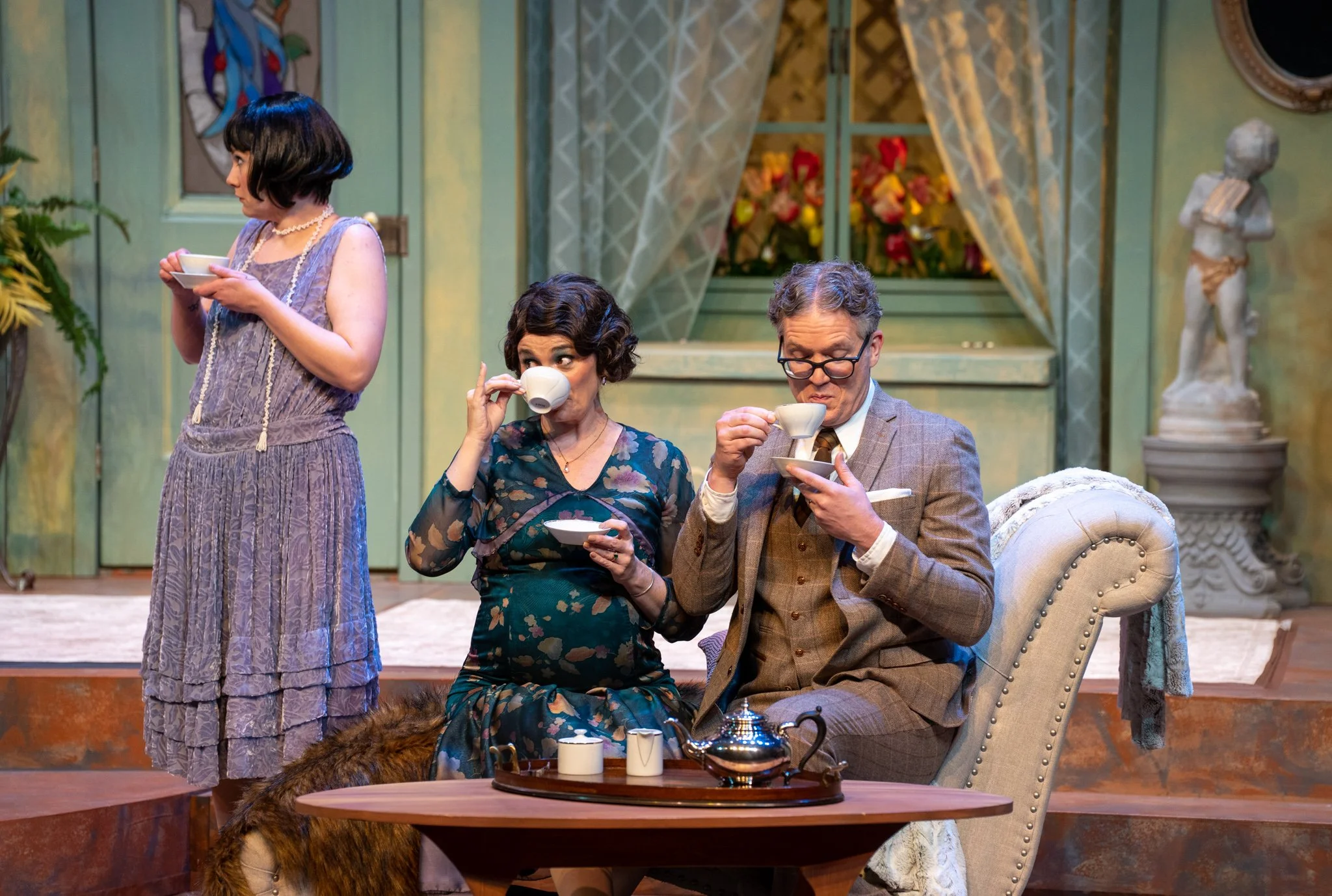 Three people having tea in a vintage living room. A woman with dark hair and wearing a patterned dress is sitting, sipping tea. A woman with curly hair in a floral dress is standing, drinking from a cup. A man with glasses in a plaid suit is seated, 