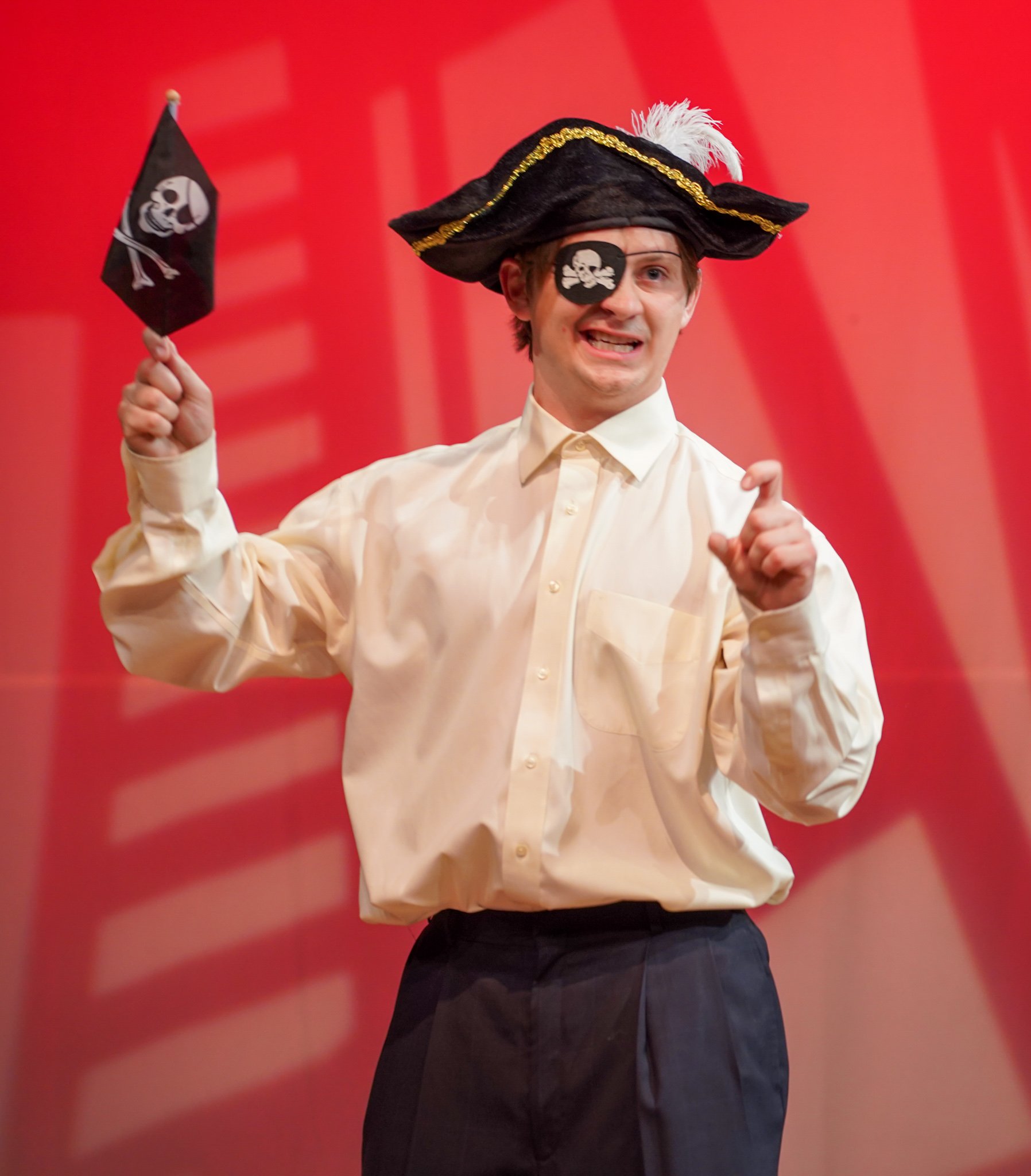 A man wearing a pirate hat with a white feather, an eyepatch with a skull and crossbones, holding a small pirate flag, and giving a thumbs-up against a red background.