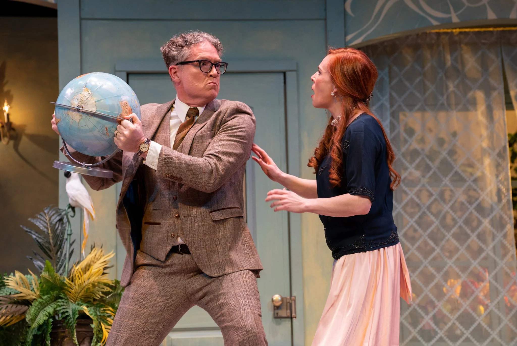 A man with glasses and a woman with red hair engaged in a heated argument indoors, with the man holding a globe and the woman reaching out with her hand.