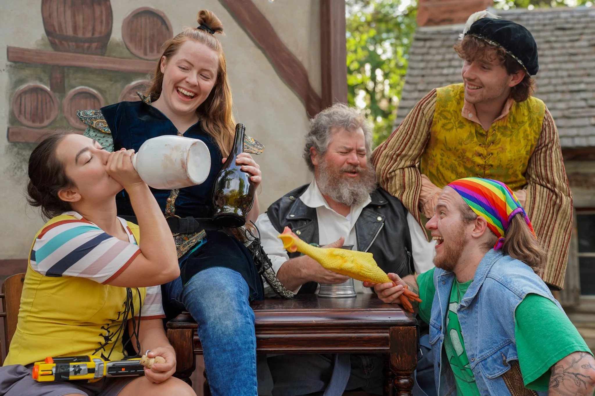A group of people in colorful costumes celebrating outdoors, with some holding beer bottles and drinking from a jug, and one person dressed as a chicken handing a chicken-shaped cake to another.