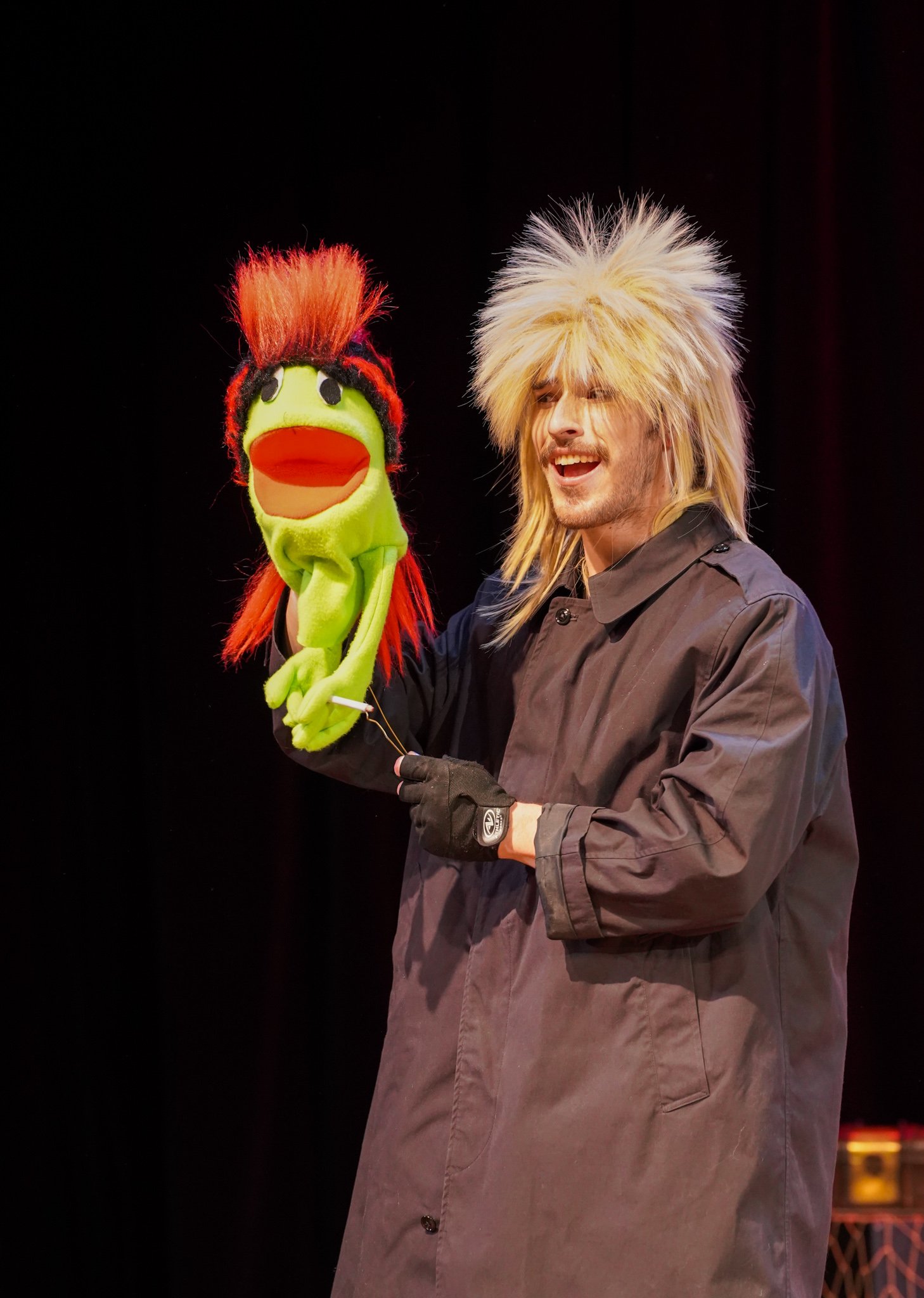 A man with a wild, blonde wig holding a puppet of a duck with a red mullet hairstyle on a black background.
