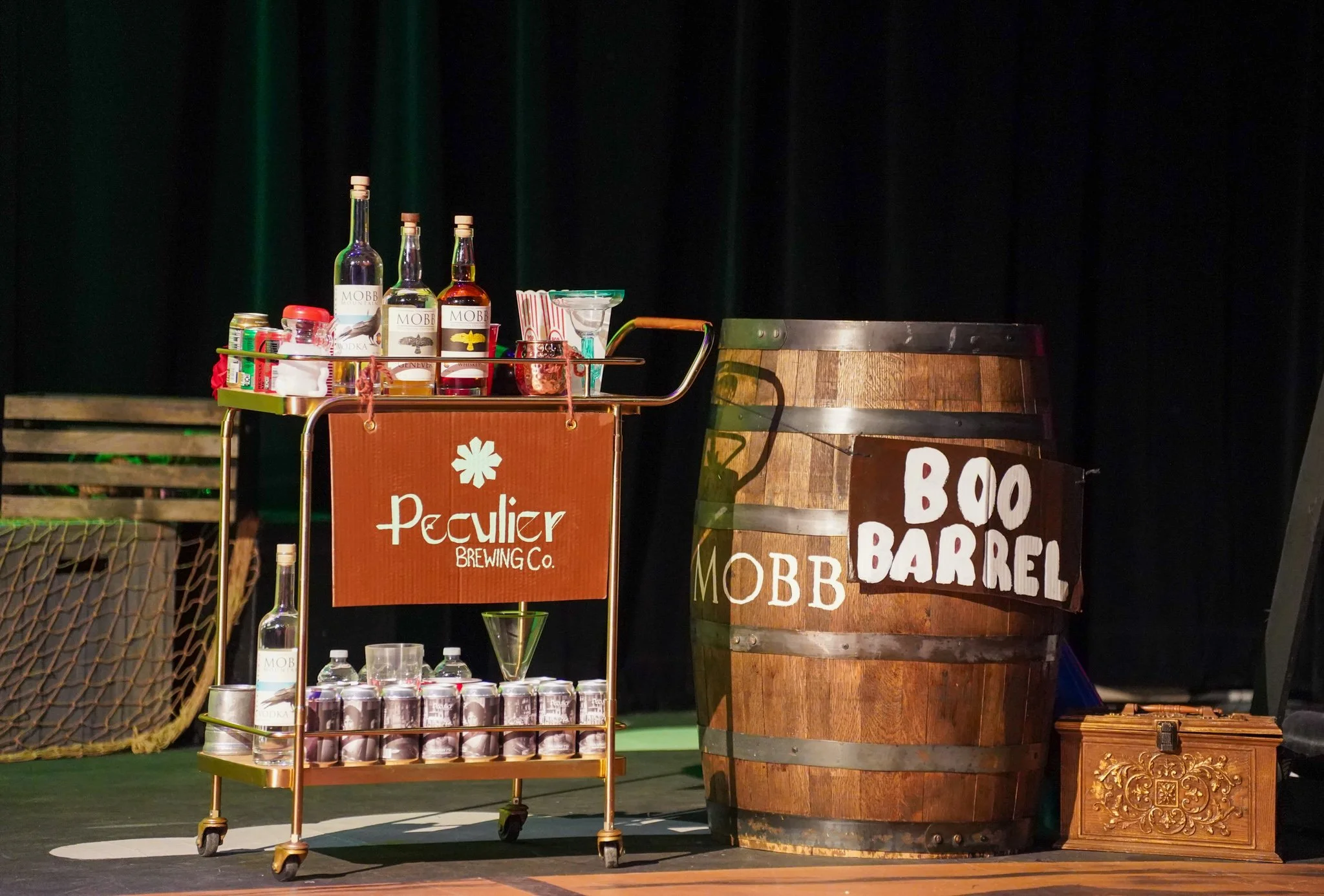Stage setup with a trolley holding bottles of vodka, cans of soda, and glasses, next to a large wooden barrel with a sign reading 'Boo Barrel' and a panel with 'Peculier Brewing Co.' logo.