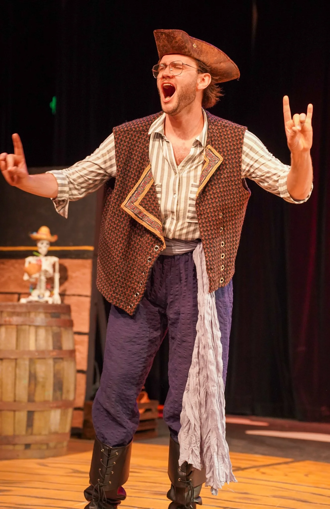 A man dressed in pirate costume singing passionately on stage, wearing a tricorn hat, glasses, a vest, baggy pants, and pirate boots, with a plastic skeleton figure in the background.