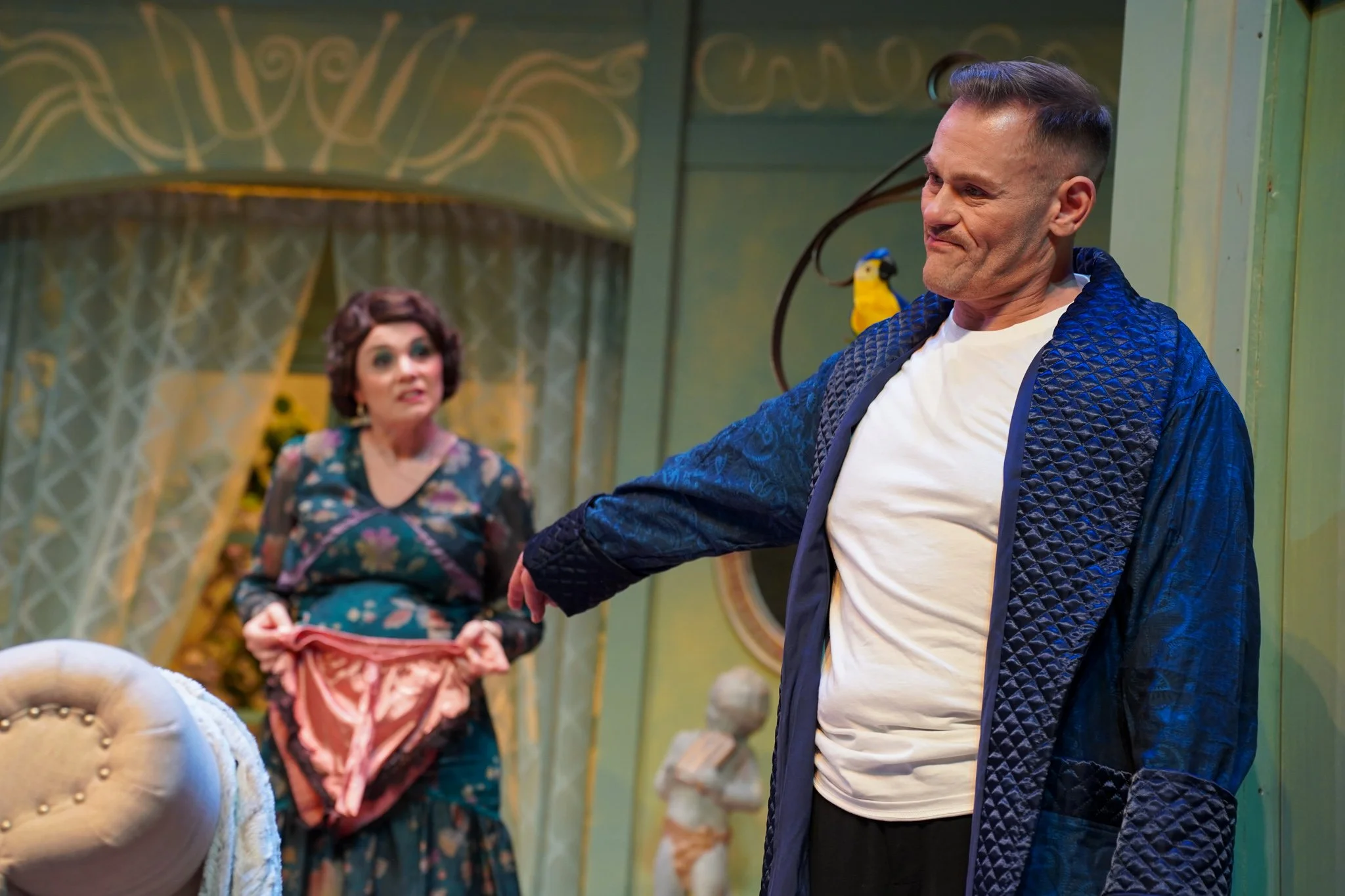 A man with short, dark hair styled upward, wearing a white shirt and a blue quilted jacket, is smiling and leaning on a piece of furniture. In the background, an elderly woman with curly, short brown hair, wearing a floral dress, is sitting and holdi