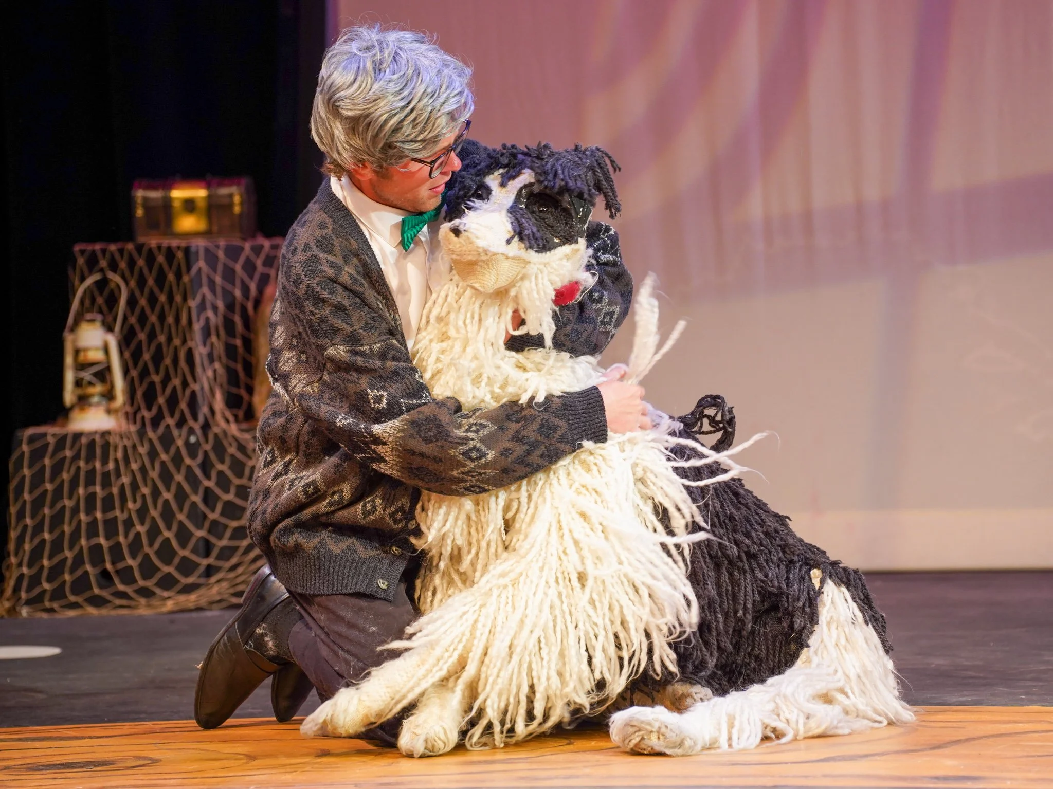 Man dressed in a suit with glasses and a green bow tie holding and cuddling a large, fuzzy puppet that resembles a dog with black, white, and purple yarn hair, on a stage with wooden flooring and a pinkish curtain.