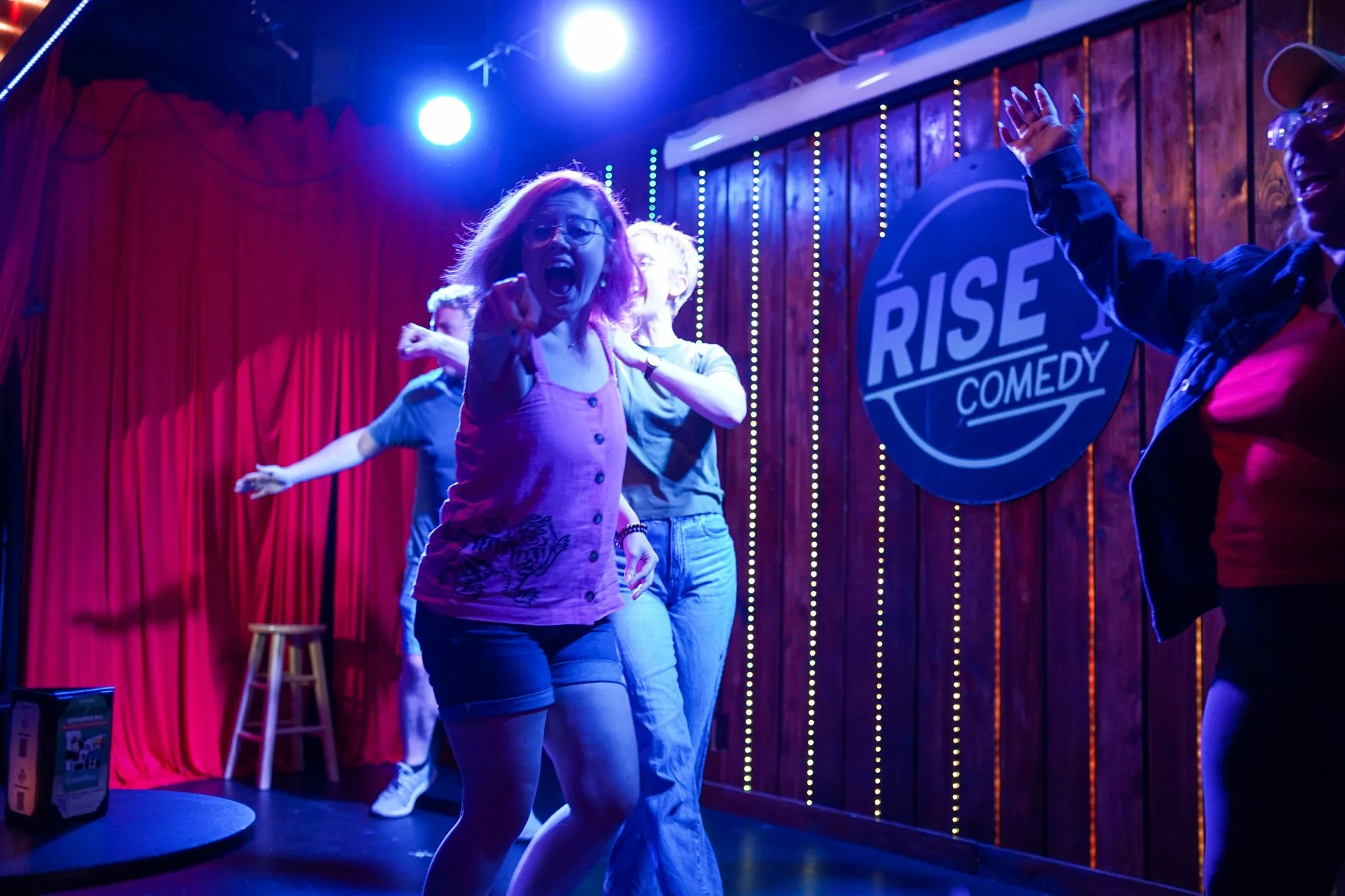 A group of people on stage at a comedy club, with red curtains and neon signs, engaging in a lively performance.