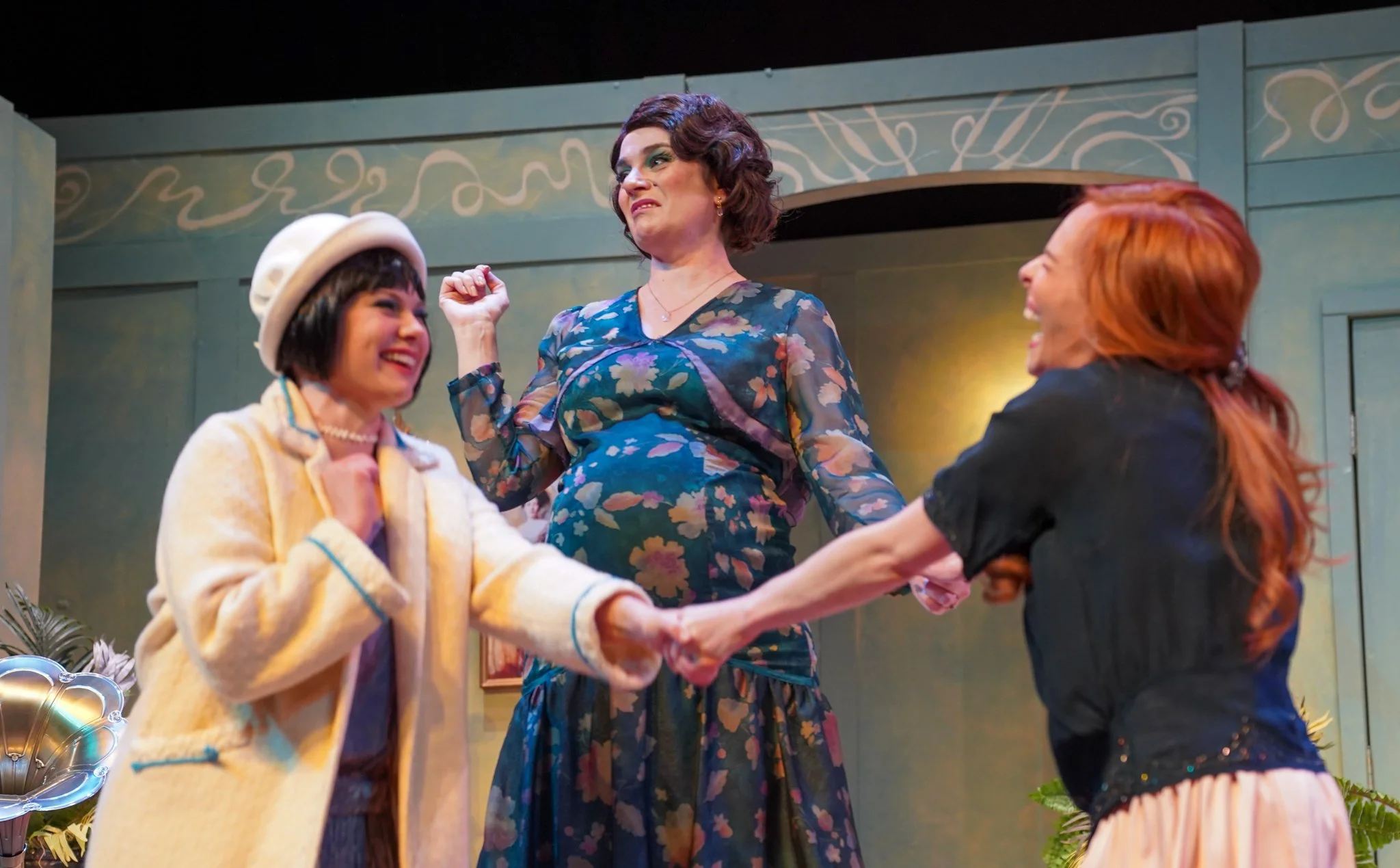 Three women on stage, two holding hands, the woman on the left wearing a cream-colored coat and white beret, the woman in the center wearing a floral dress and gesturing with her right hand, and the woman on the right with red hair laughing. There ar
