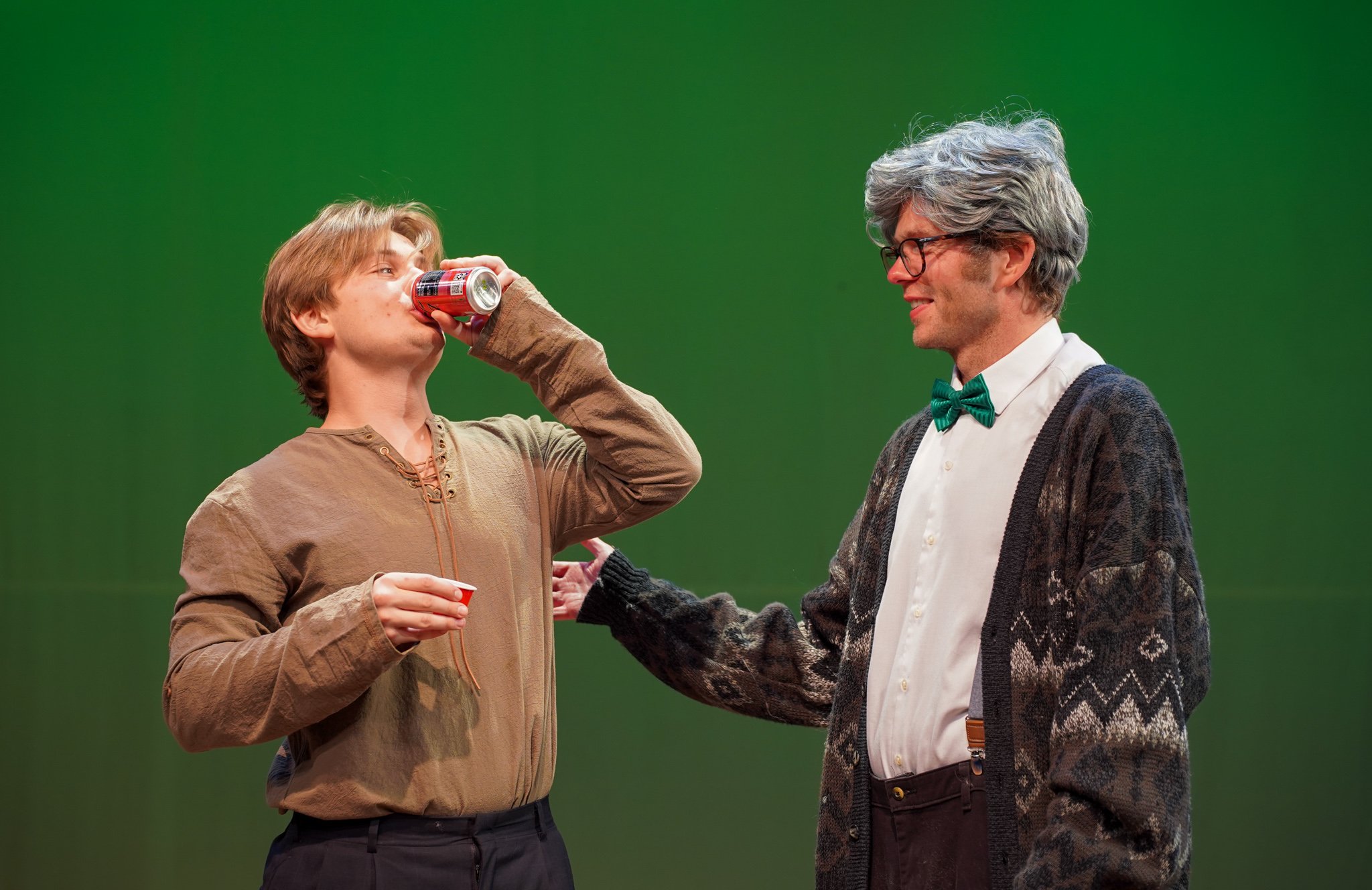 A young man with blond hair is drinking from a soda can while holding a small red cup in his left hand. An older man with gray hair and glasses, wearing a white shirt, a green bow tie, and a patterned cardigan, is smiling at him and touching his arm.