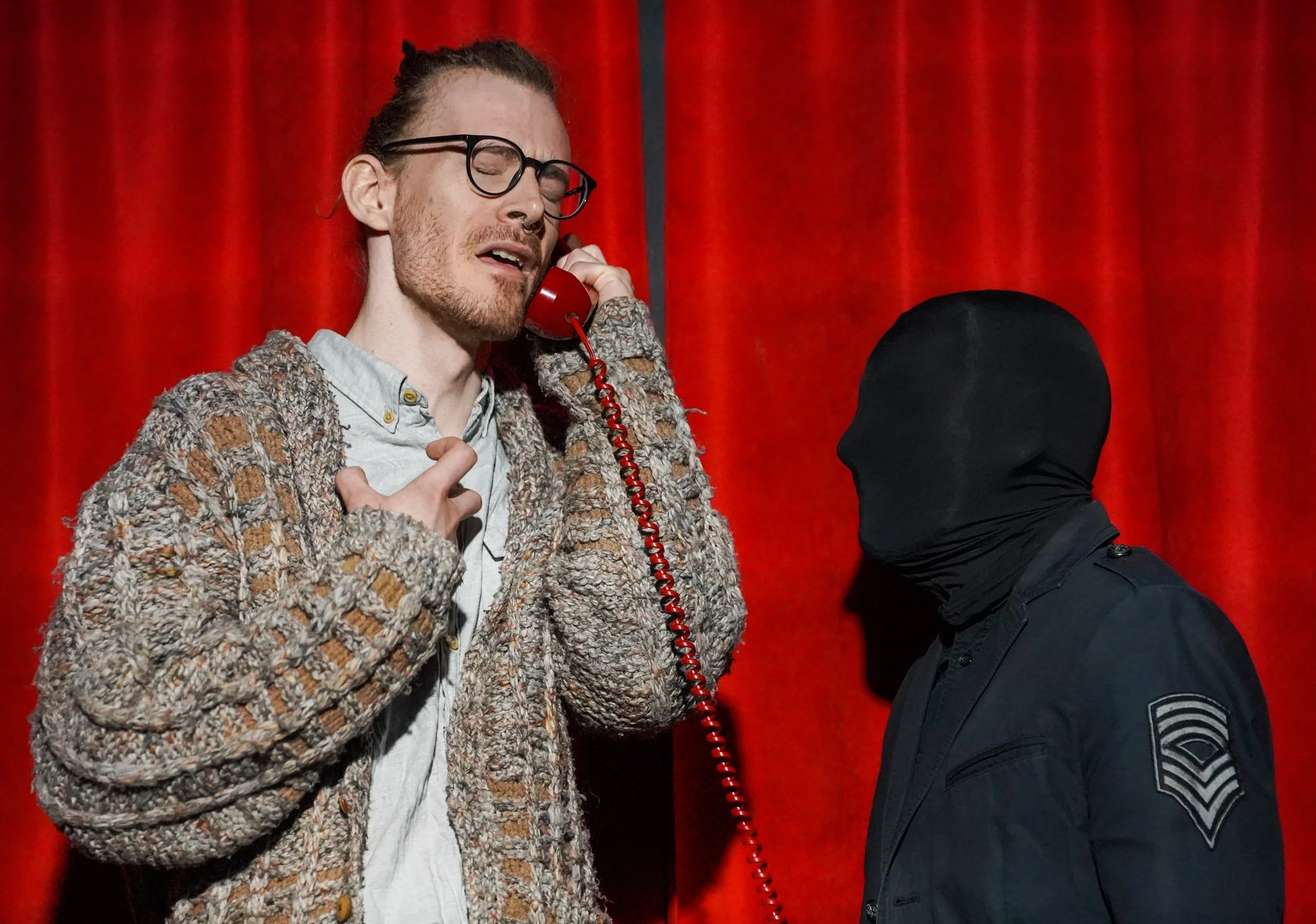 A man with glasses, a beard, and a nose ring holding a red telephone receiver, with a person in a black hooded outfit standing next to him against a red curtain background.