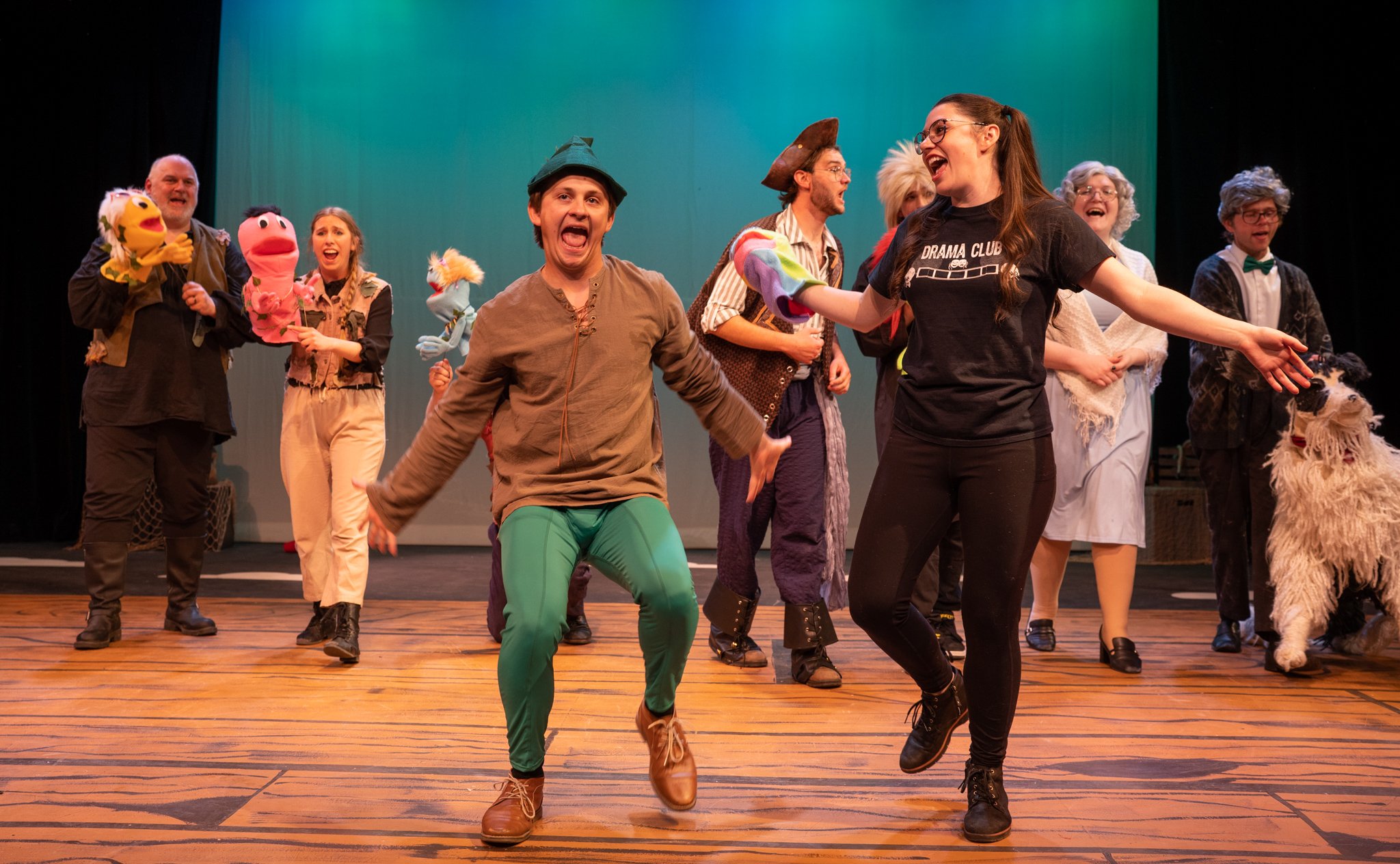 The stage is filled with performers, some holding puppets, wearing costumes, and singing or dancing, in a lively theatrical production.