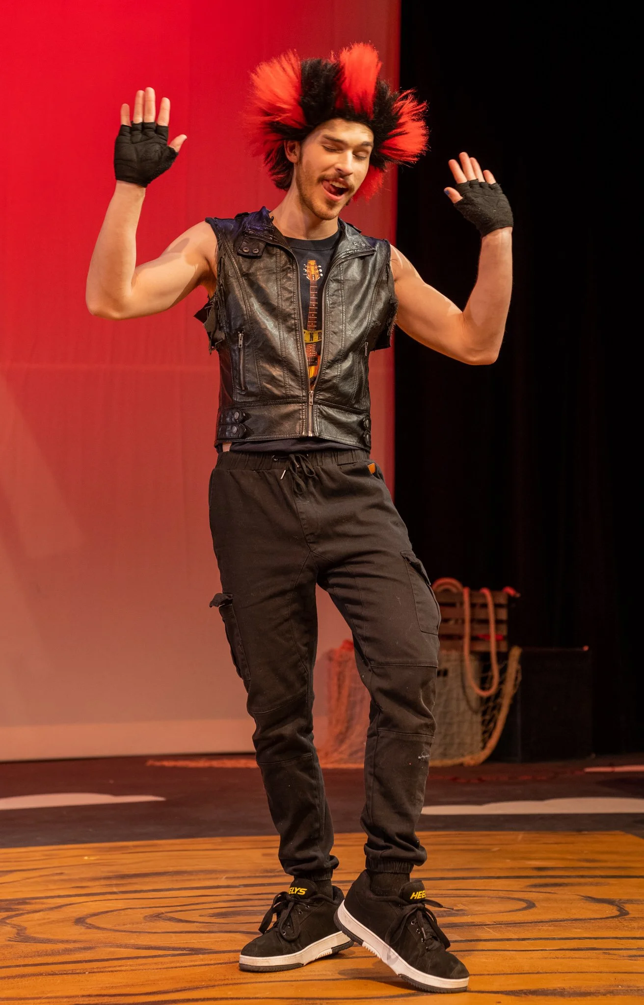 A man with a multicolored hairstyle, wearing a sleeveless leather vest, black pants, and black sneakers, dancing on a wooden stage with red and black background.