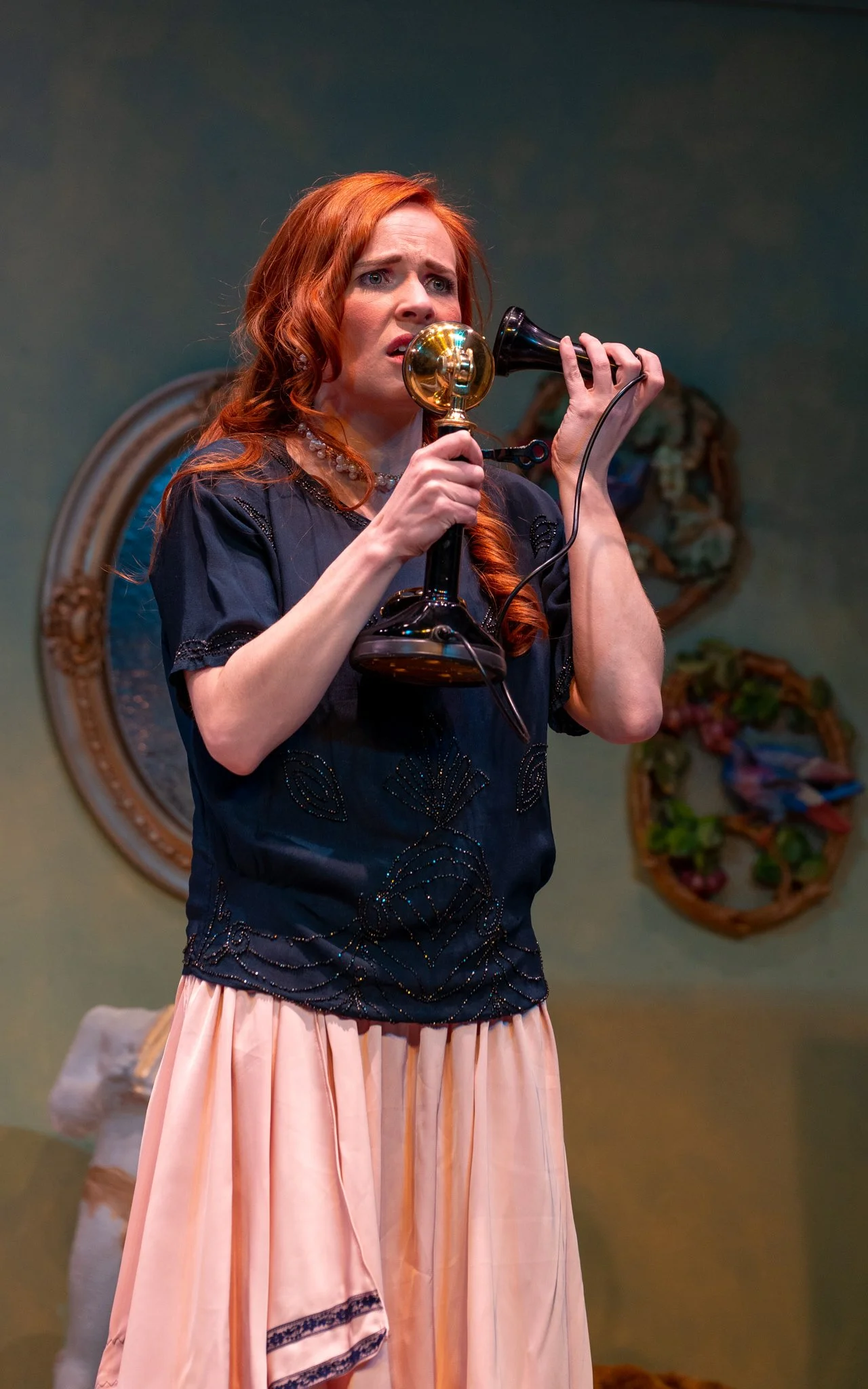 A woman with long red hair is holding an old-fashioned black and gold telephone to her ear, looking concerned or worried. She is wearing a dark blue embroidered blouse and a peach-colored skirt, standing in a room with a green wall. Two decorative wr