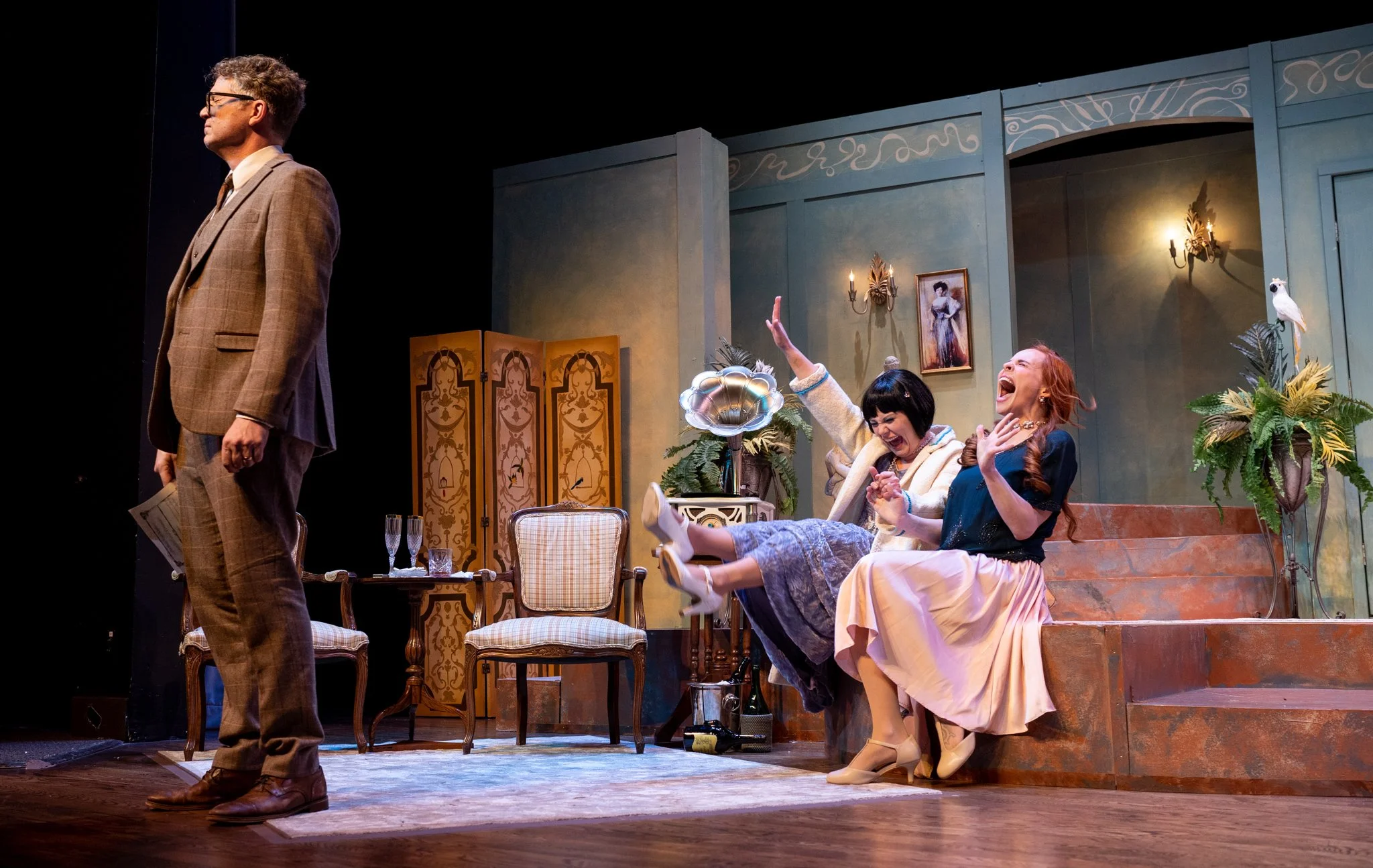 A theatrical stage scene with four actors, one man standing on the left with glasses and a plaid suit, three women seated and laughing on the right, with vintage decor, plants, and paintings in the background.