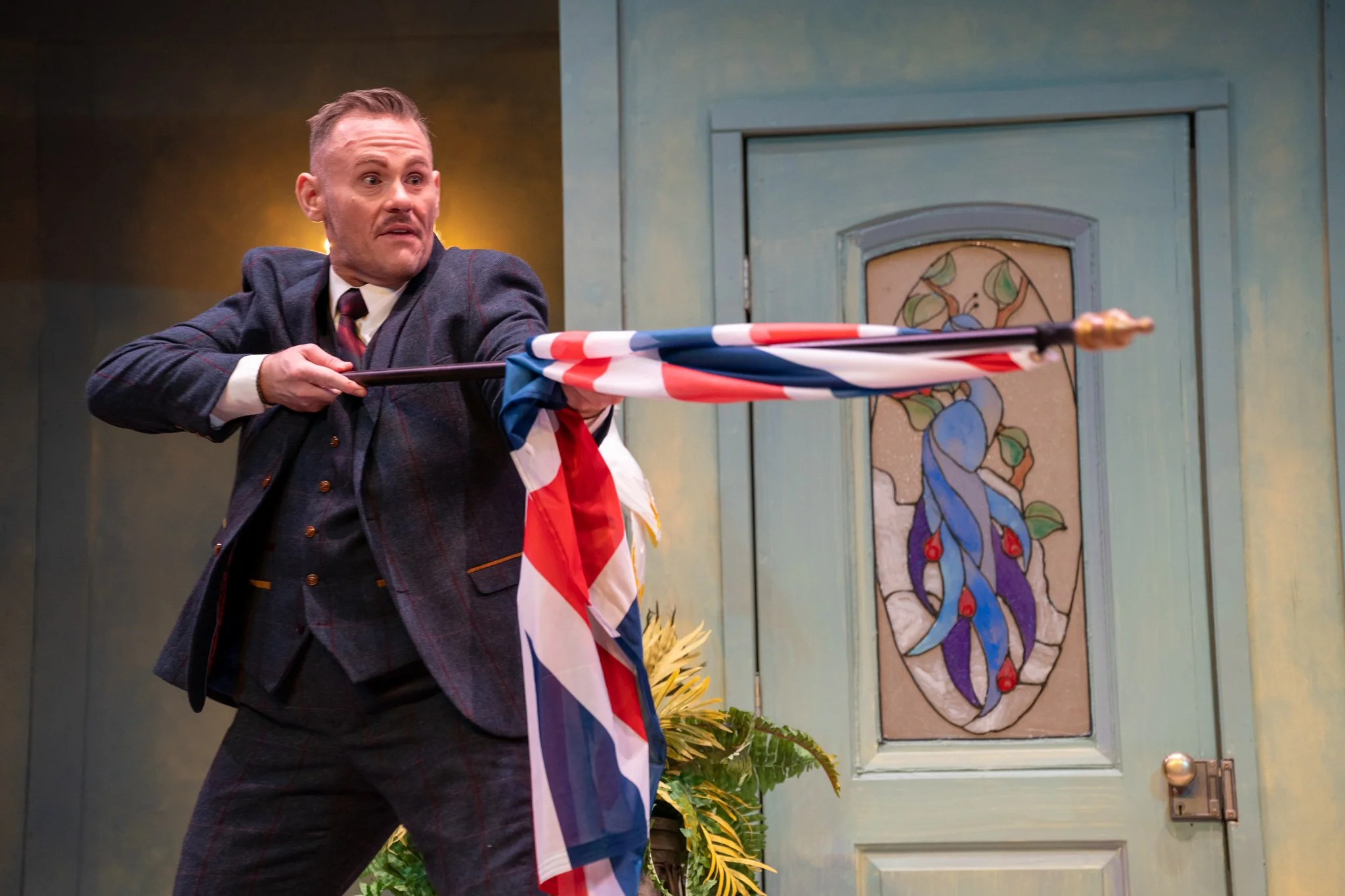 A man in a plaid suit and white shirt with a tie holding an umbrella with a red, white, and blue pattern in a room with blue walls and decorative doors. The man appears surprised or shocked.
