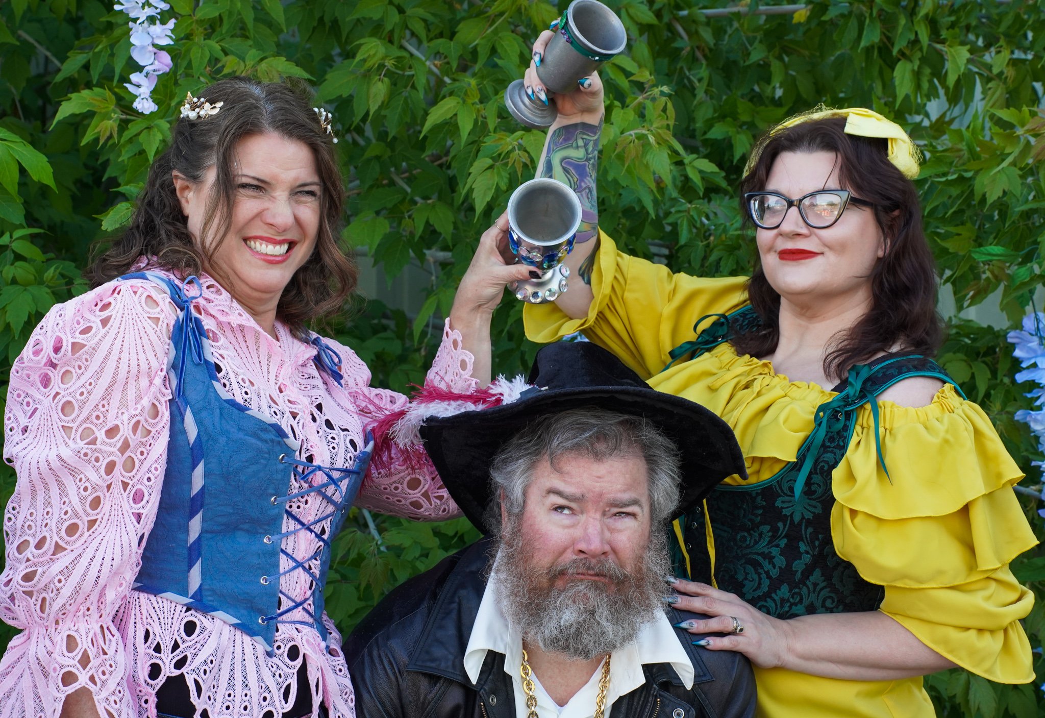 Two women dressed in colorful, playful costumes hold goblets in a celebratory pose behind a man with a gray beard wearing a hat and costume, outdoors with green leafy trees in the background.