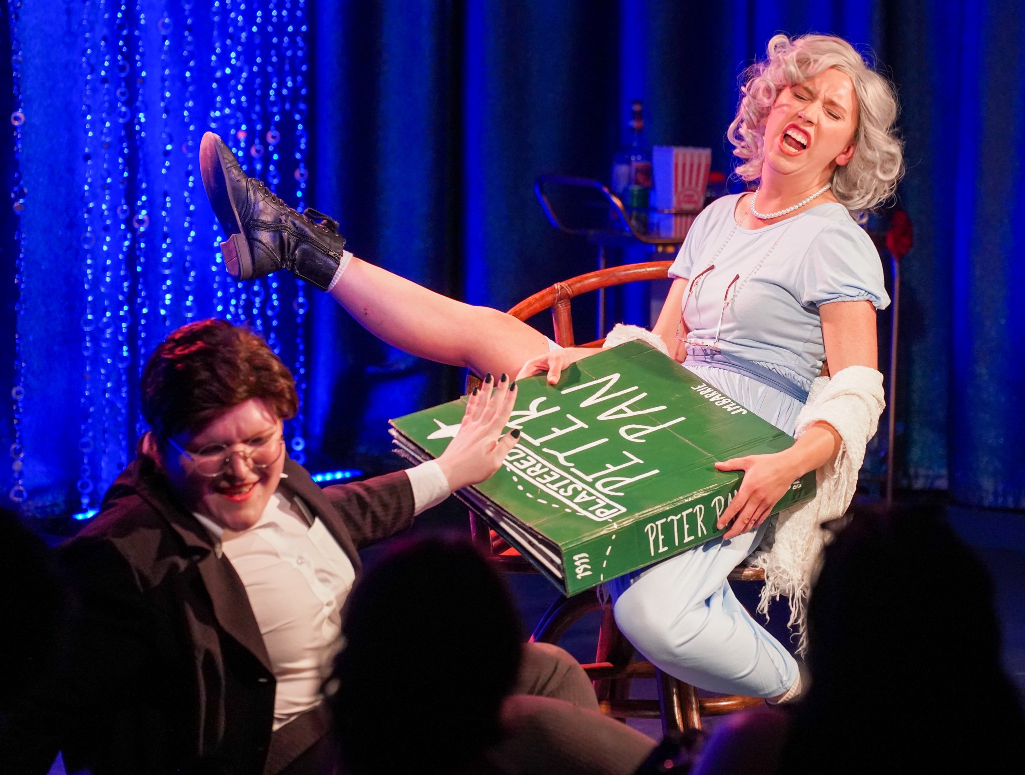 A woman with curly blonde hair singing on stage, with her leg raised on a chair, wearing a light blue dress, pearl necklace, and black boots, while holding a large green and white sign. Two other people are sitting in front of her, one in a dark blaz