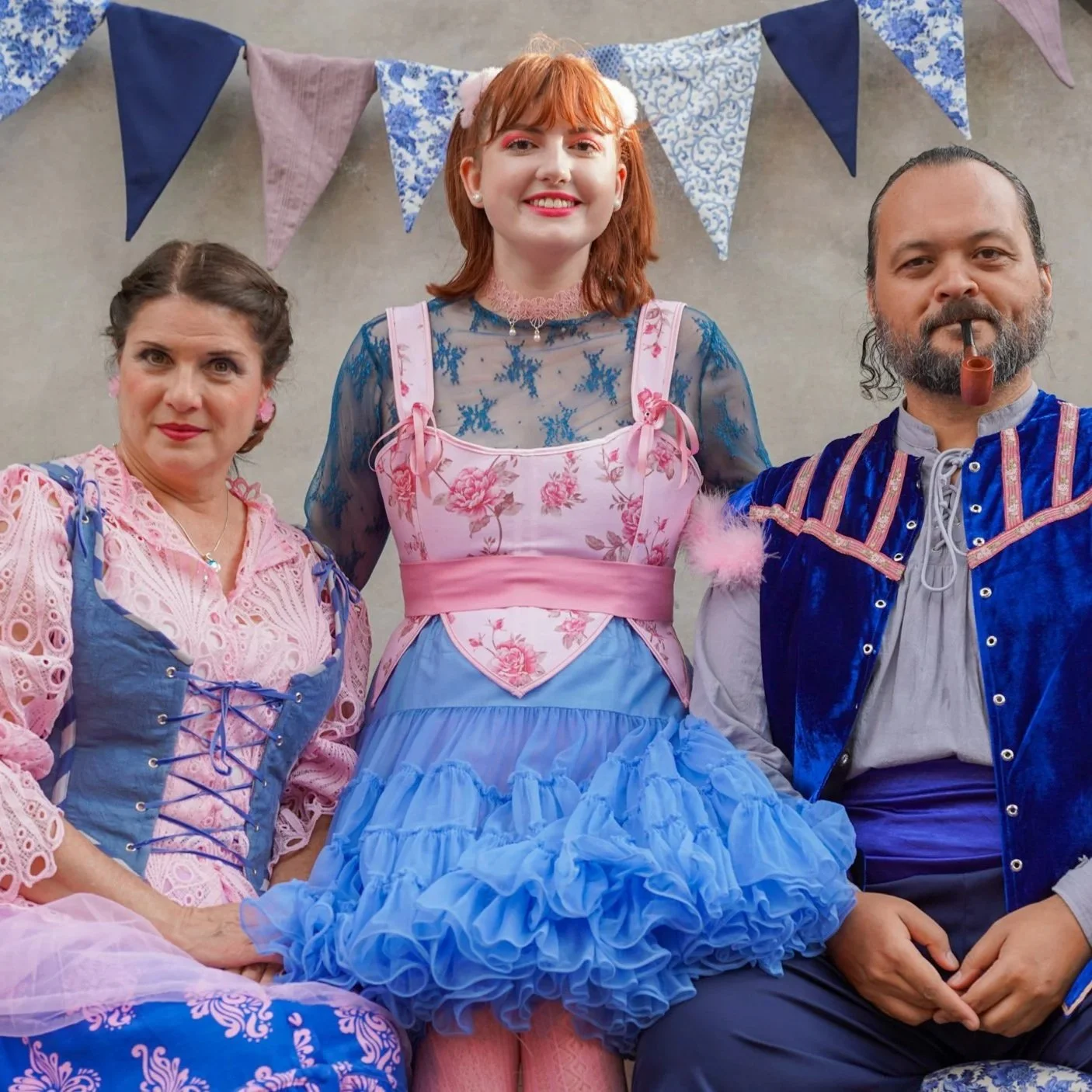 Three people dressed in colorful, vintage-style costumes pose for a photo. The woman in the center wears a blue and pink floral dress with a ruffled blue skirt, the woman on the left wears a pink and blue lace dress, and the man on the right wears a blue velvet jacket with pink accents and a gray shirt. There are blue and purple bunting flags hanging behind them.