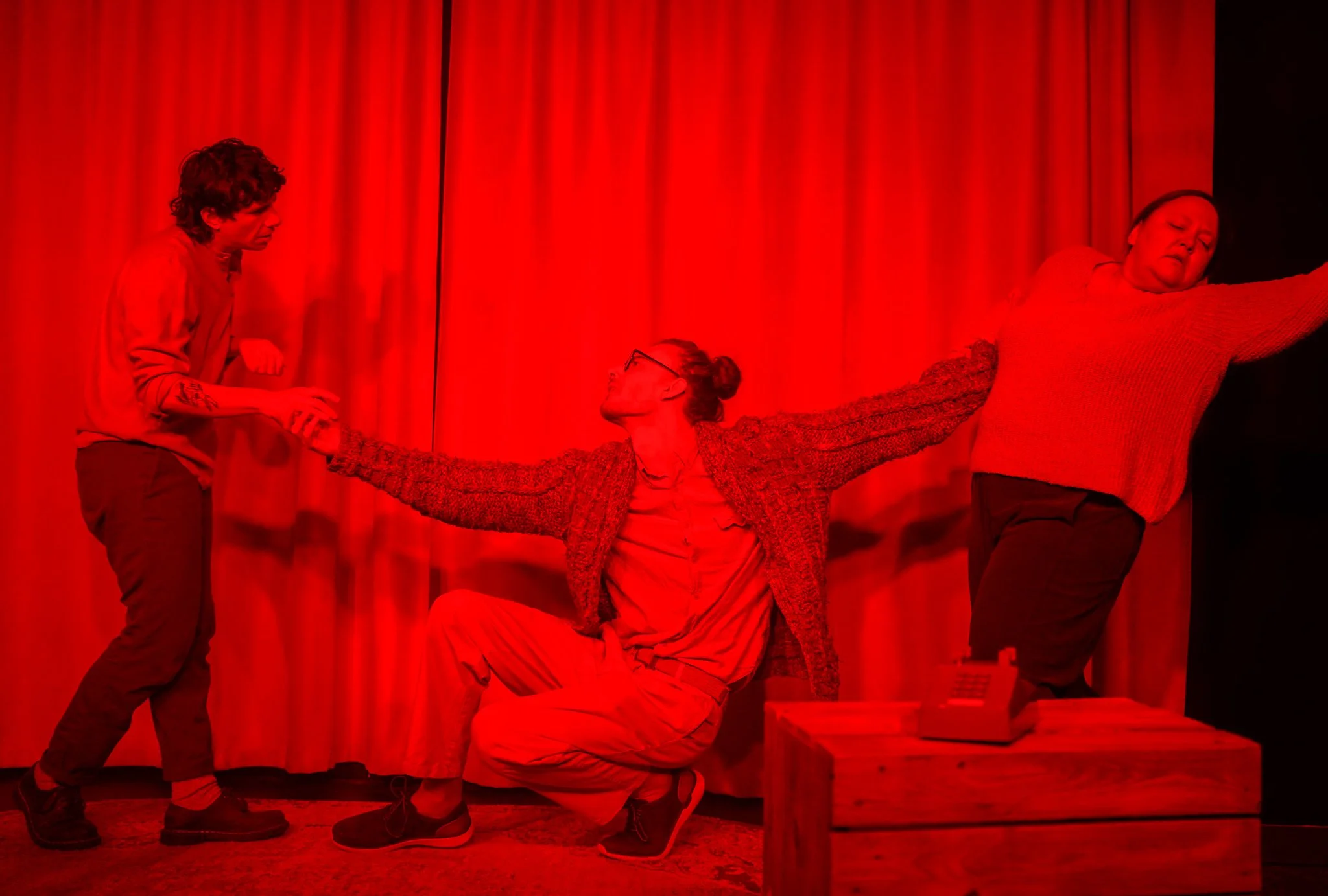 Three people on stage with red curtains in the background. One person is crouched with arms outstretched, another person is standing with a serious expression, and a third person is standing with arms extended outward.