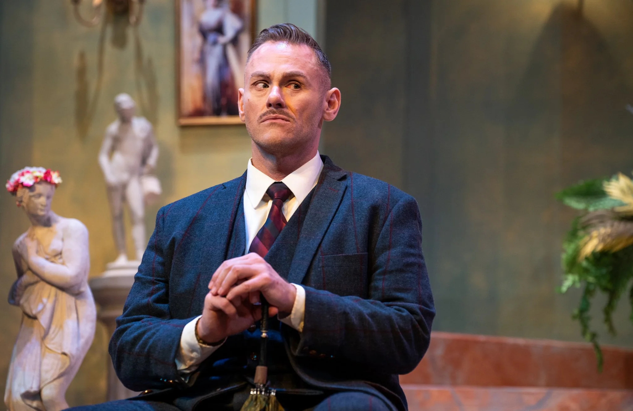 A man with slicked-back hair, wearing a checkered suit and bow tie, sits with his hands clasped in front of him, exhibiting a skeptical or disapproving expression. The background features classical sculptures and art, suggesting a museum or art galle