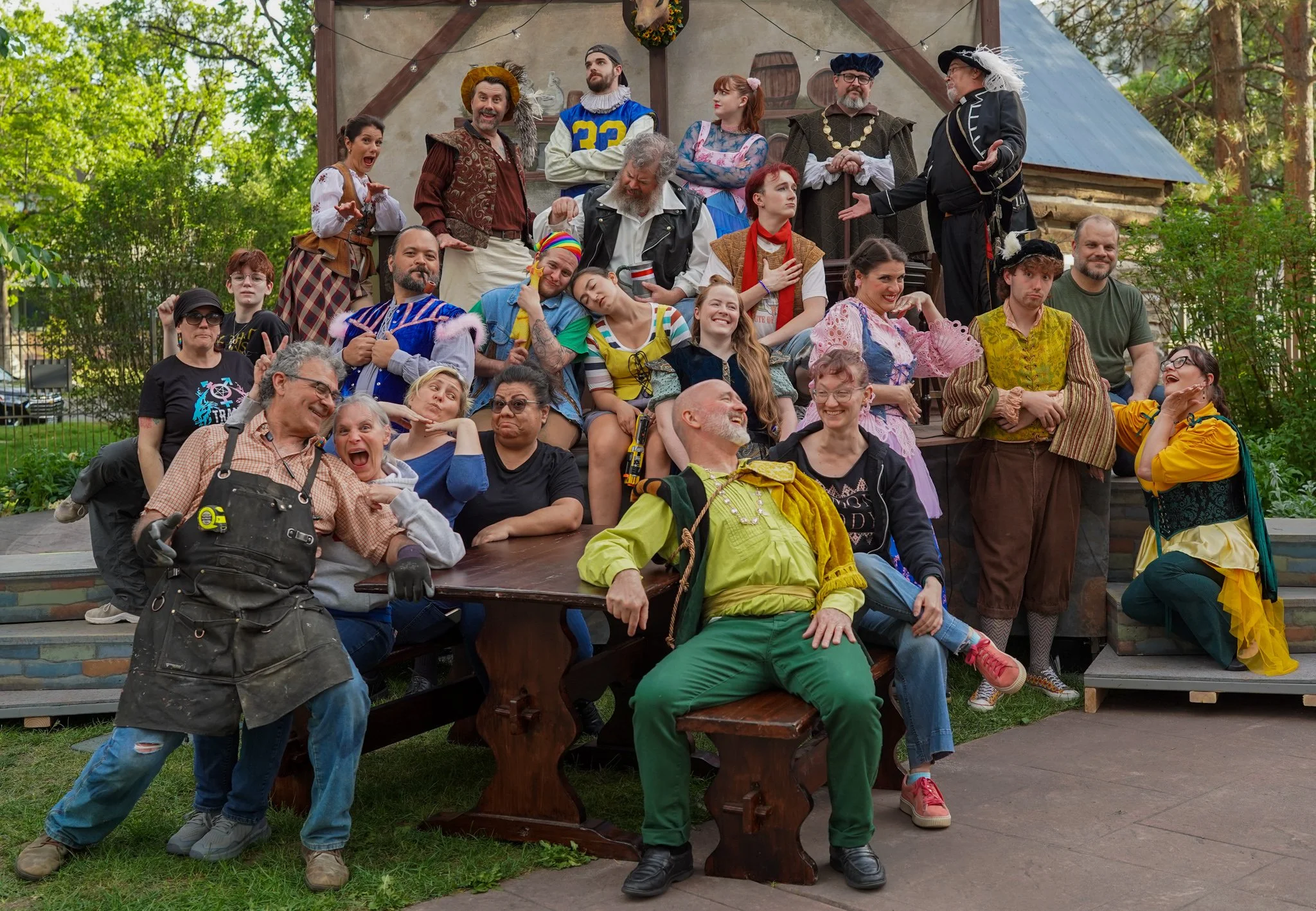 A group of people in colorful costumes and casual clothing posing on and around a wooden table and steps outdoors, with a backdrop of trees and a rustic building.