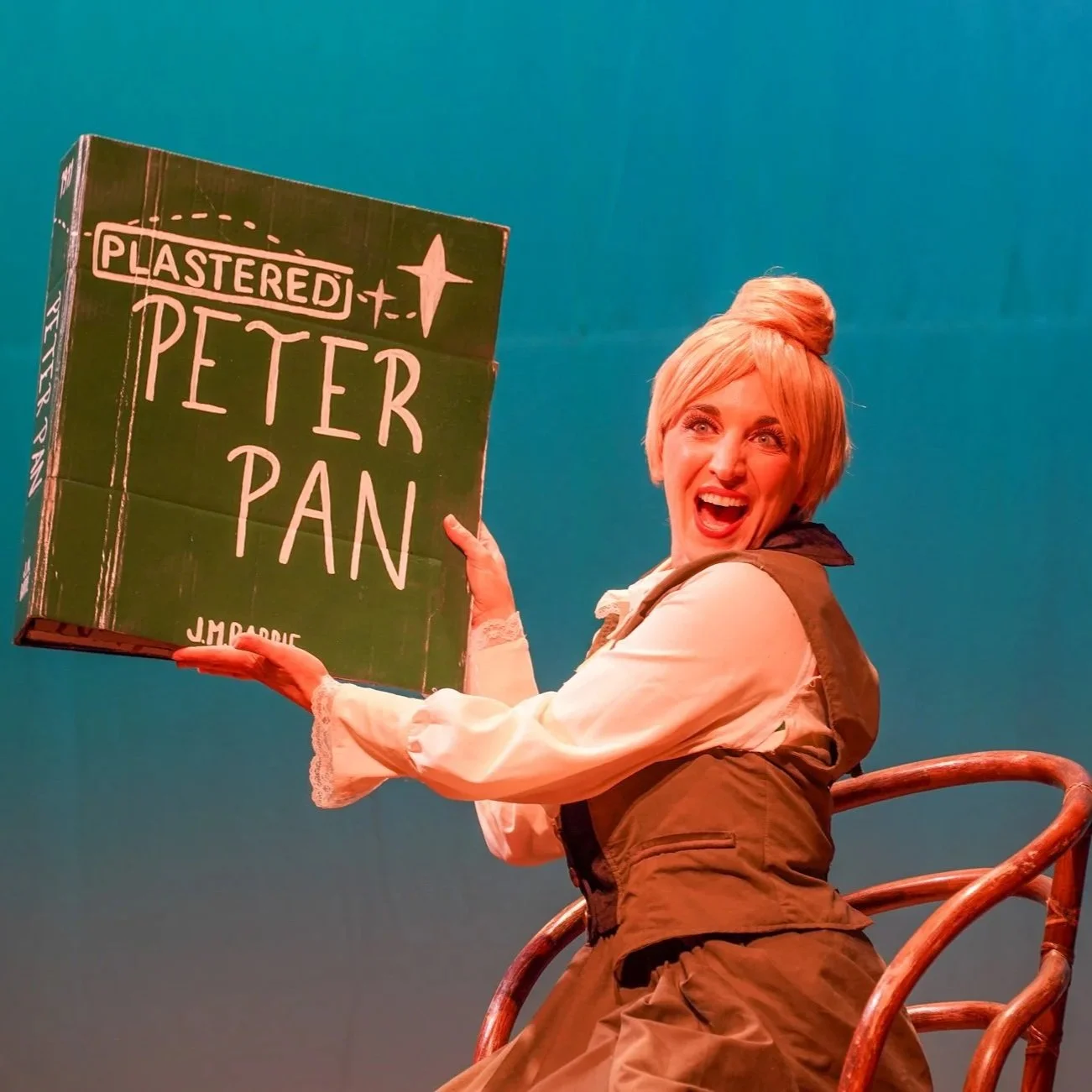 A woman with blonde hair styled in a bun, wearing period costume, holding a box labeled "Plastered Peter Pan" on a theatrical stage with a teal background.
