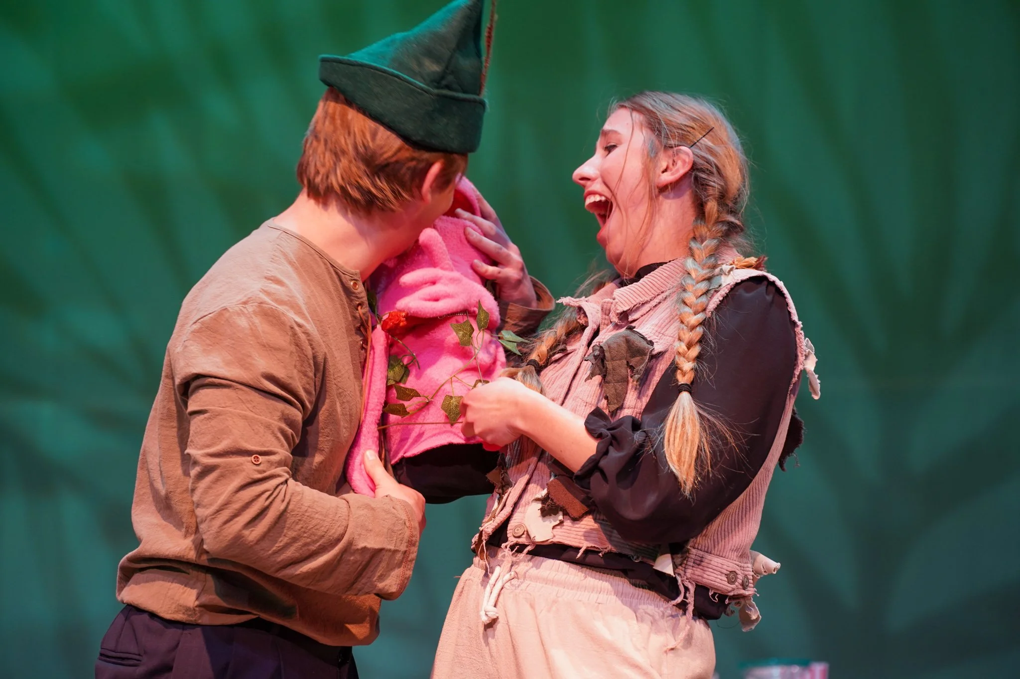 A man and woman share a joyful moment on stage, with the man facing away, wearing a brown shirt and green hat, and the woman smiling brightly with braids, wearing a patchwork-style vest and skirt, holding a small pink stuffed animal.
