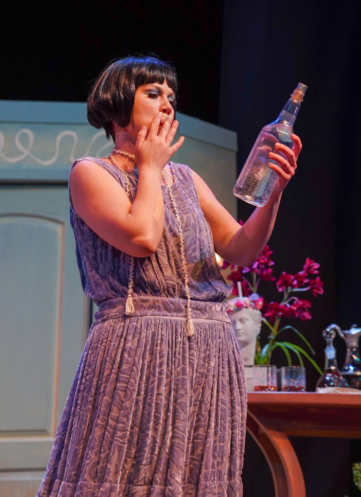 Woman with short dark hair in a dress looking at a clear bottle, in a room decorated with pink flowers and a bust sculpture.