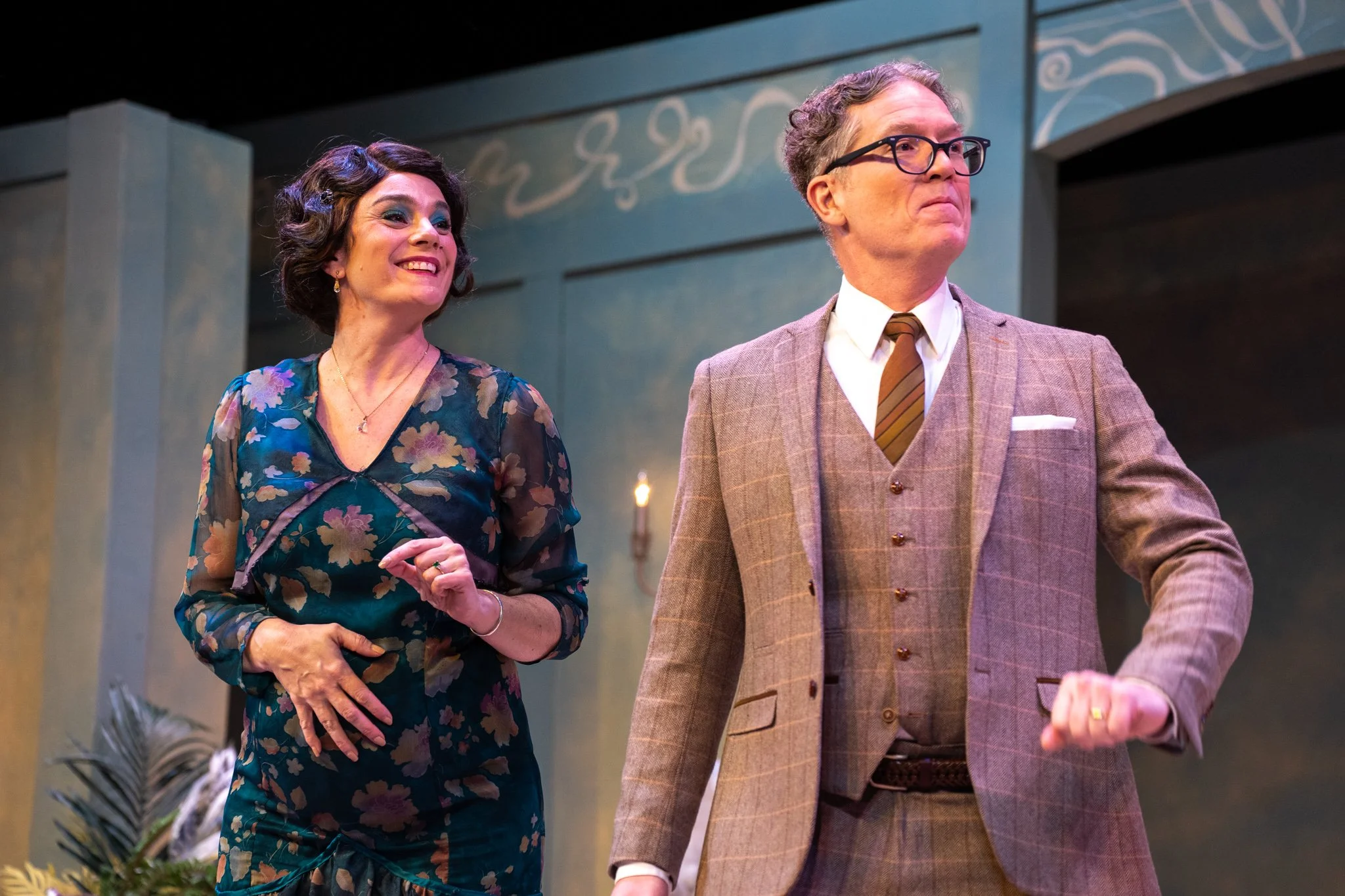 A woman in a floral dress smiling and a man in a brown checked suit with glasses, both standing indoors with a decorated background. 