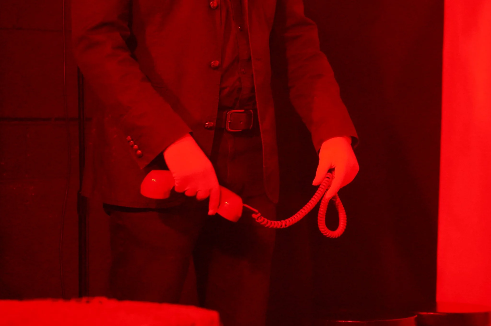 Person holding a red landline telephone with curly cord in red-lit room, wearing a jacket and black belt.
