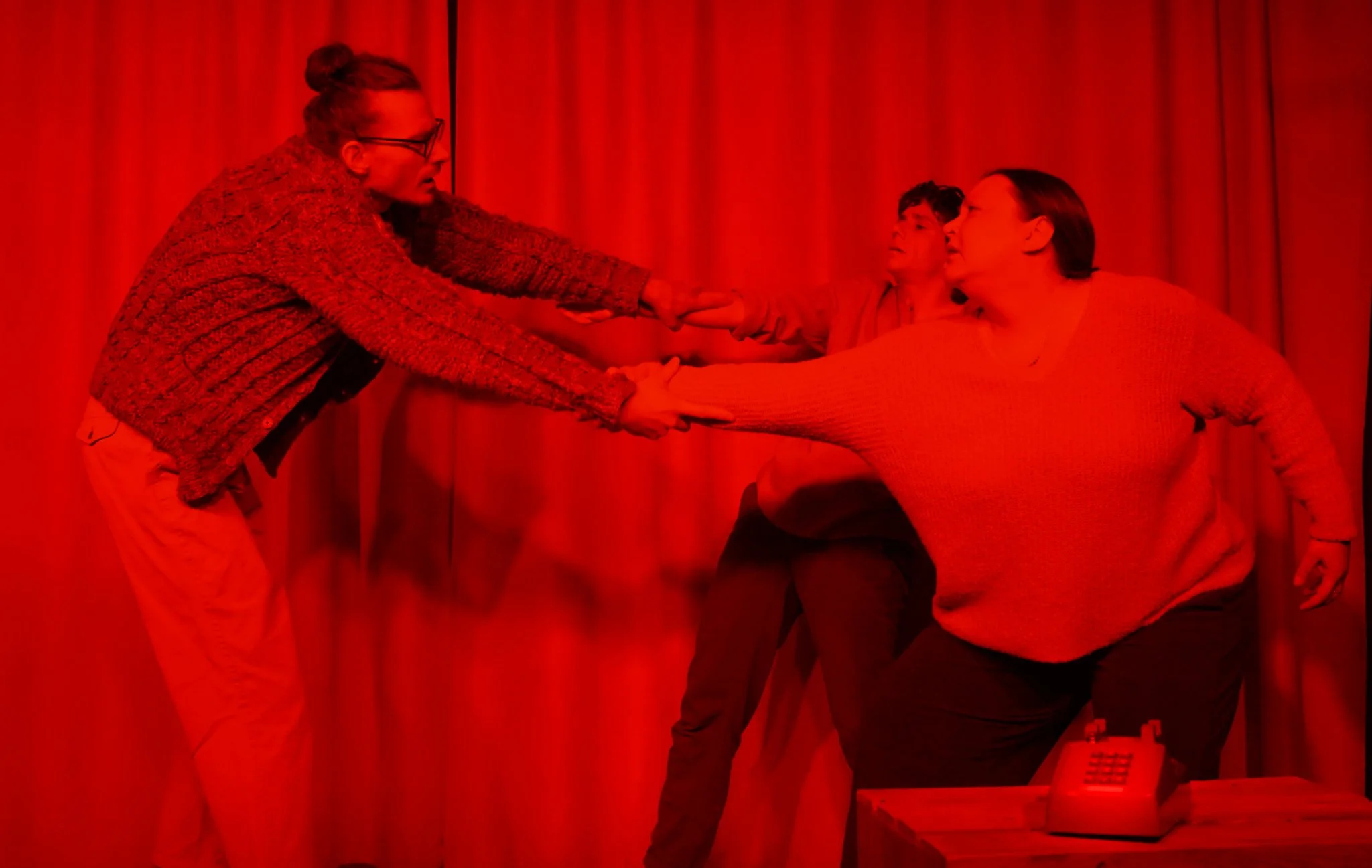 Three women having a heated debate behind a red curtain. The woman on the left is wearing glasses and a multicolored sweater, holding hands with the woman on the right who is wearing a red sweater. A woman in the middle looks surprised or concerned. 